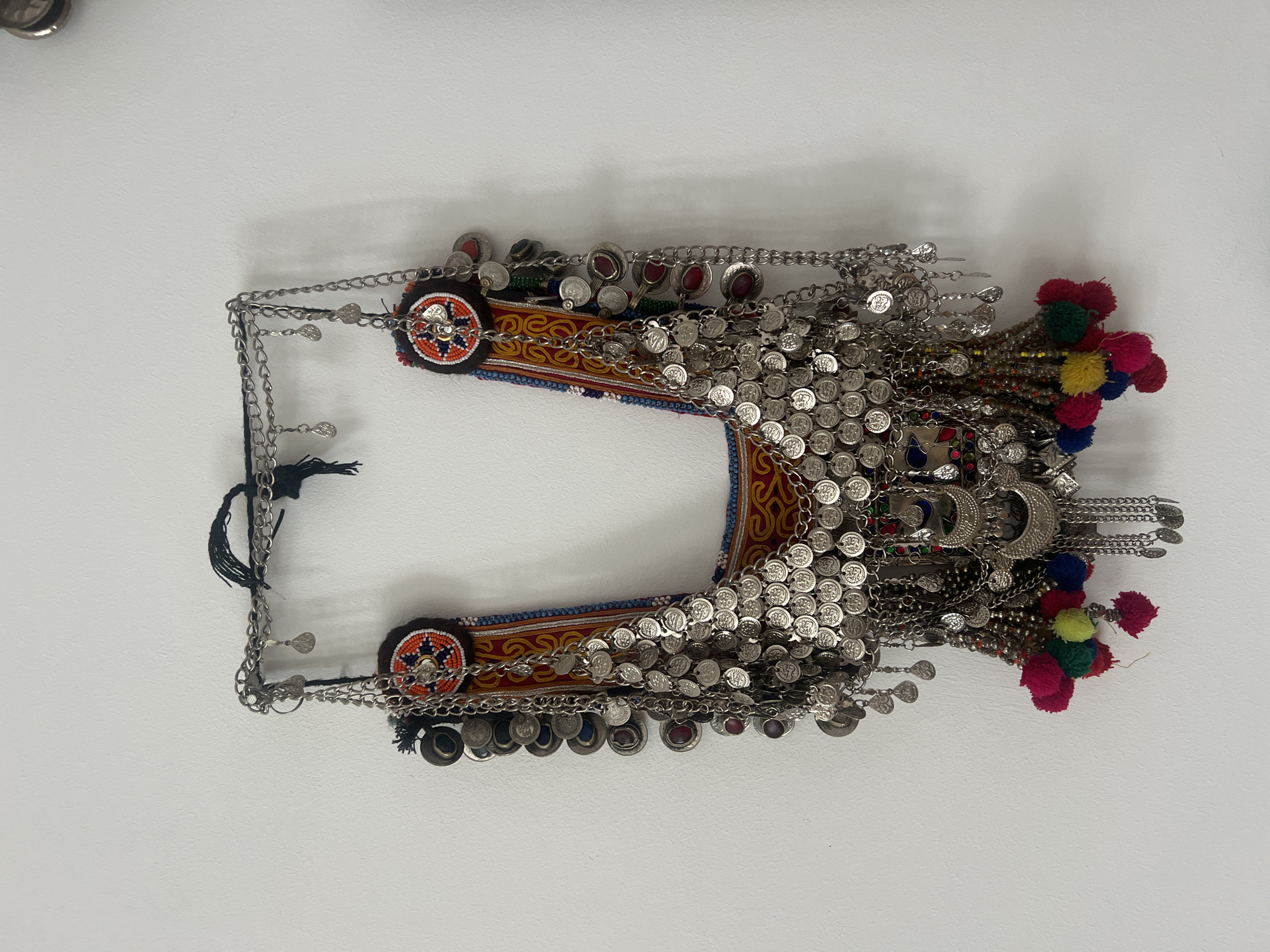 The image shows a detailed view of a traditional tribal headdress or breastplate lying flat against a white surface. The central component is an ornate fabric piece with colorful embroidery, predominantly featuring red, yellow, and blue patterns. Affixed to this fabric are numerous silver-colored metal discs or coins, densely arranged and hanging as adornments. The headdress also incorporates beaded sections, small stones, and decorative chains. On each end of the fabric there are circular medallions and tassels in various colors. There is no indication this item is located in Berlin, Germany.