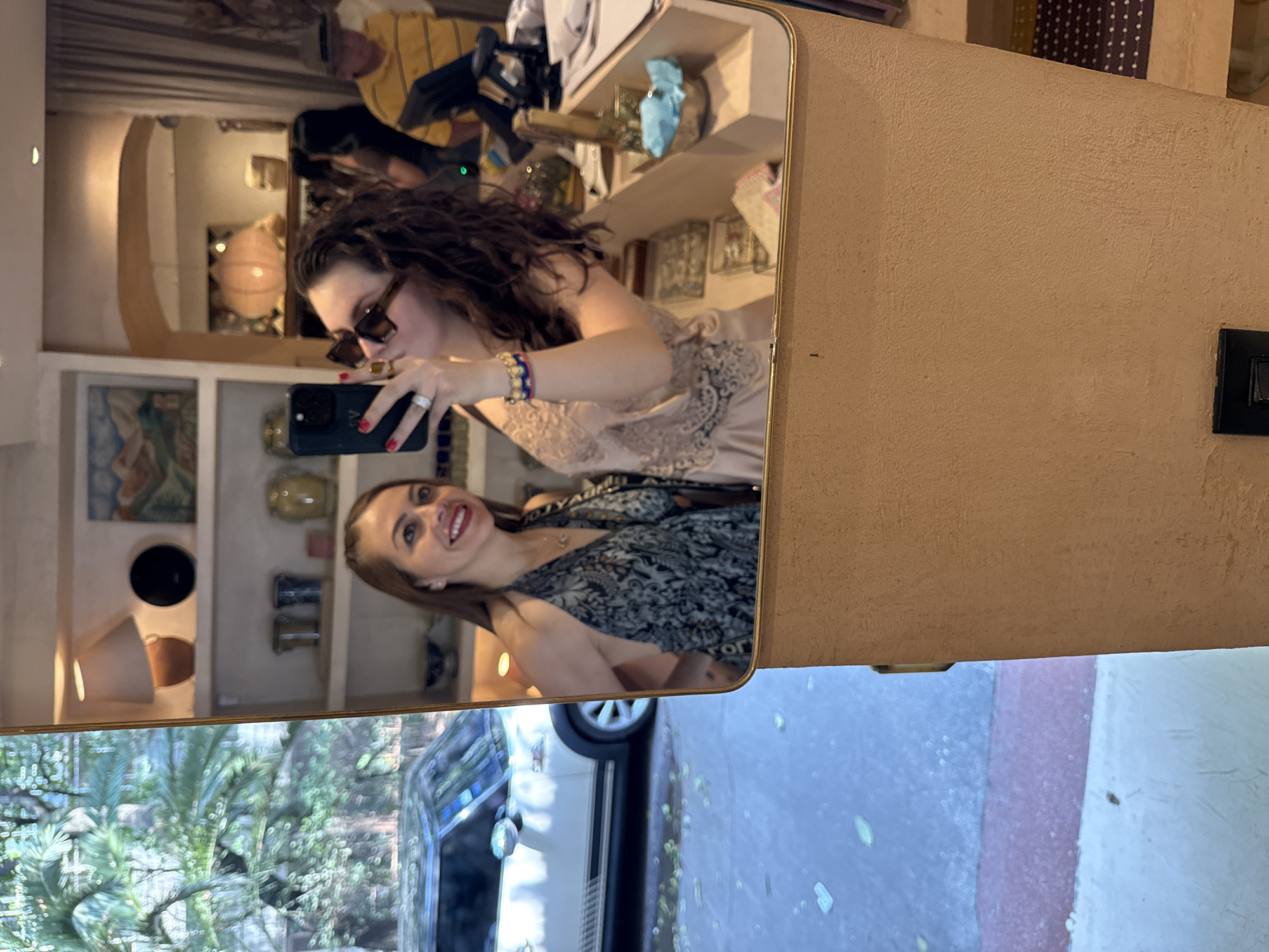 A vertical selfie taken in a mirror captures two women inside what appears to be a cafe or shop in Mexico City, Mexico.
In the foreground of the mirror's reflection, a woman with dark, curly hair and rectangular sunglasses holds a black smartphone, seemingly taking the picture. She wears a light beige or peach-colored top with lace detailing and has a blue and red beaded bracelet on her left wrist, along with a silver ring.
Beside her reflection, another woman with light brown hair and a bright smile looks towards the camera. She wears a dark top or dress with a light-colored, intricate pattern, possibly with the letters "OR" visible, suggesting a brand. A delicate necklace is visible around her neck.
The background of the mirror's reflection reveals an inviting indoor setting with light-colored walls and shelving displaying various decorative items, including pottery, paintings, and a spherical light fixture. A man in a yellow and black striped shirt is partially reflected further in the background.
Outside the window or glass door, a lush green landscape with palm-like foliage is visible, along with a white car (possibly a Mini Cooper) parked on what looks like a street or sidewalk featuring painted red and white stripes. The bright outdoor light suggests it is daytime. On the right side of the image, outside the mirror, a textured beige wall with a black light switch is visible.