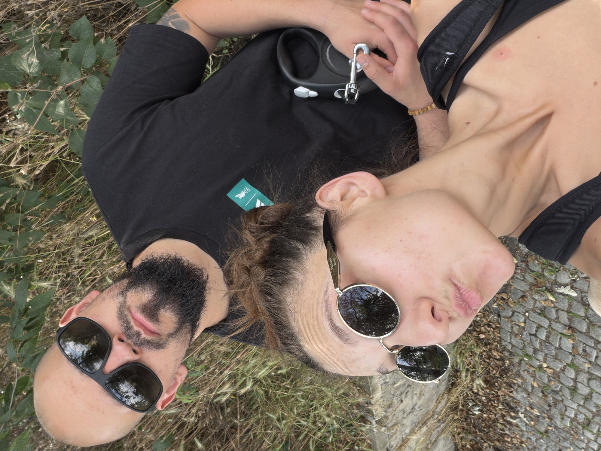 The image captures two individuals in an outdoor setting within Plovdiv, Bulgaria. A bald man with a dark beard and black rectangular sunglasses is positioned behind and slightly above the woman. He wears a black t-shirt featuring a green bird logo, resembling the Liverpool FC crest, on the left chest and displays a dark tattoo on his left upper arm. He holds a black retractable dog leash in his right hand. In the foreground, a woman with light brown hair tied in a bun is taking a selfie, making a puckered lip expression. She wears gold-framed round sunglasses with "PRADA MILANO" visible on the right arm. Her attire includes a black tank top and black backpack straps. A small, circular, reddish mark is visible on her upper right chest, and she wears a light-colored beaded bracelet on her right wrist. The surrounding environment consists of dry grass, green leafy vegetation, and a cobblestone ground visible on the lower left.
