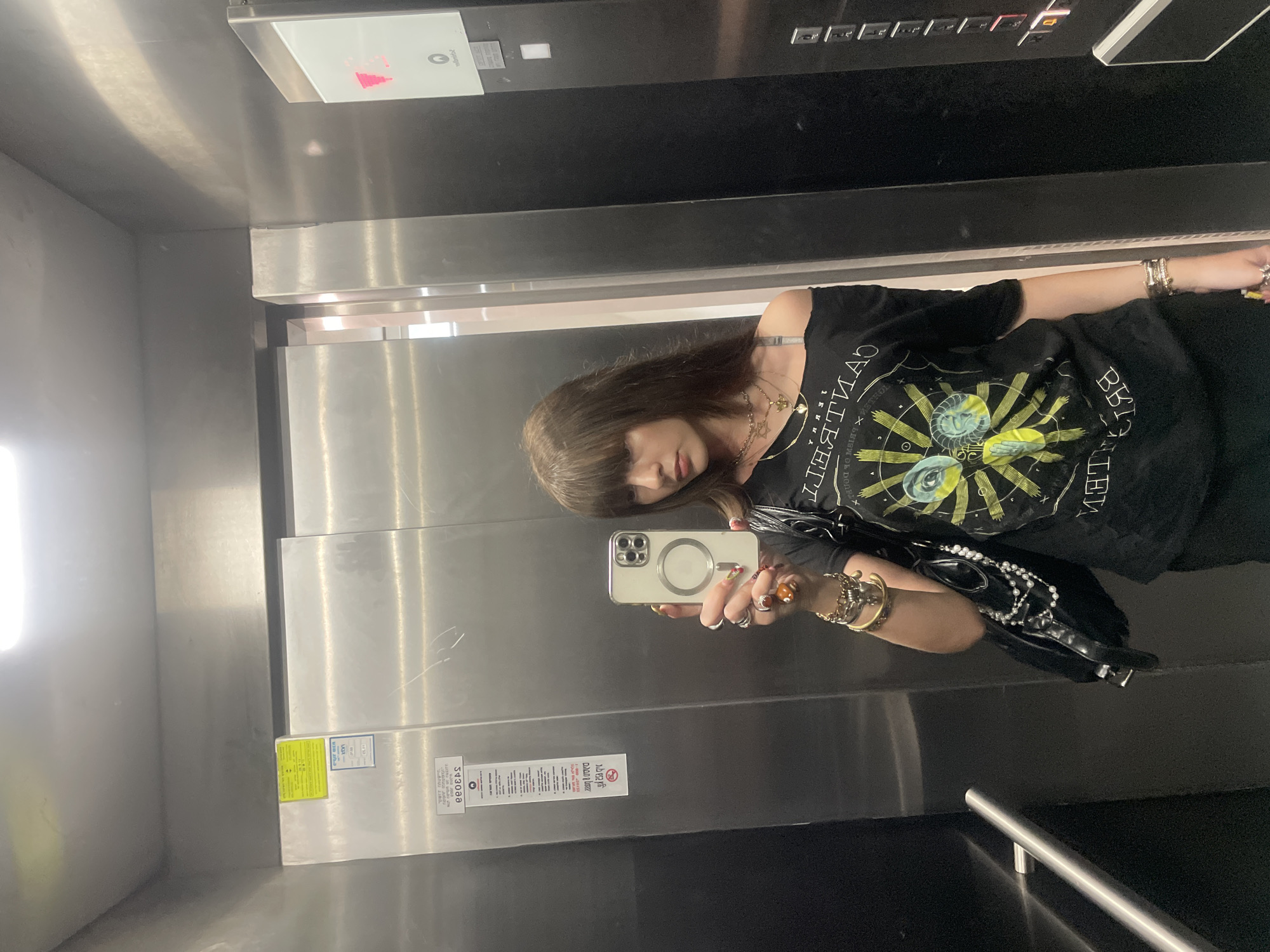 A woman is taking a selfie in what appears to be an elevator. The interior of the elevator is predominantly metal, with a control panel visible at the top of the frame. The woman is holding a smartphone with a MagSafe charger ring showing on the back. She is wearing a black off-shoulder t-shirt with graphic designs. Accessories include necklaces, bracelets, and rings. Part of a black purse is visible at the right of the frame. Based on the informational markings on the elevator panel, it is likely located in Raanana, Israel.