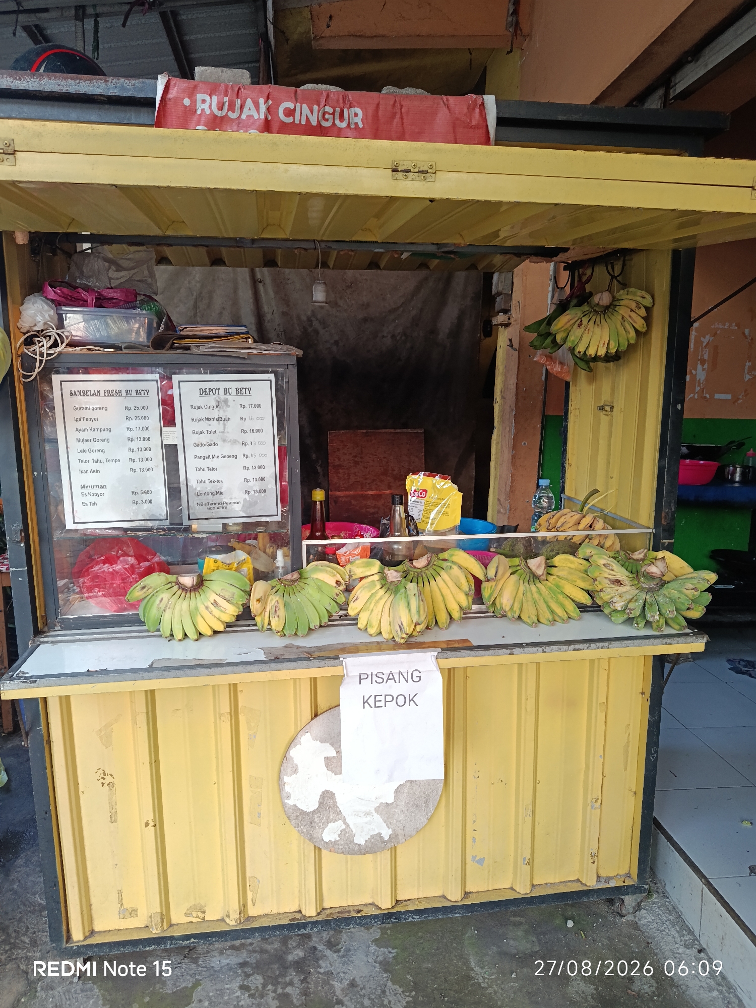 The image shows an outdoor food stall in Keboansikep, Indonesia. The stall is yellow and has a red banner with "RUJAK CINGUR" written on it. Inside the stall, there are two menu boards, one titled "SAMBELAN FRESH BU BETTY" and the other "DEPOT BU BETTY". Both boards list various food items and their prices. In front of the stall, there are several bunches of bananas displayed on a counter. A white sign with "PISANG KEPOK" is also visible. The ground in front of the stall appears to be wet. The overall scene suggests a typical street food vendor in Indonesia.
