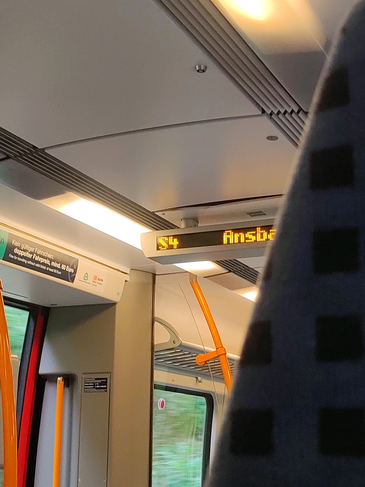 The image shows the interior of a train. A digital display shows "S4" and "Ansb". A poster on the left side of the train is partially visible, with German text that says "Kein gültiger Fahrschein: doppelter Fahrpreis, mind. 60 Euro". The train is in motion, as indicated by the blurred scenery outside the window. The destination display likely indicates a route number and the next stop. Based on this information and external knowledge, this train is likely on the S4 line heading towards Ansbach. Roßtal is a municipality in the district of Fürth, Bavaria, Germany, and it is served by the S4 line.