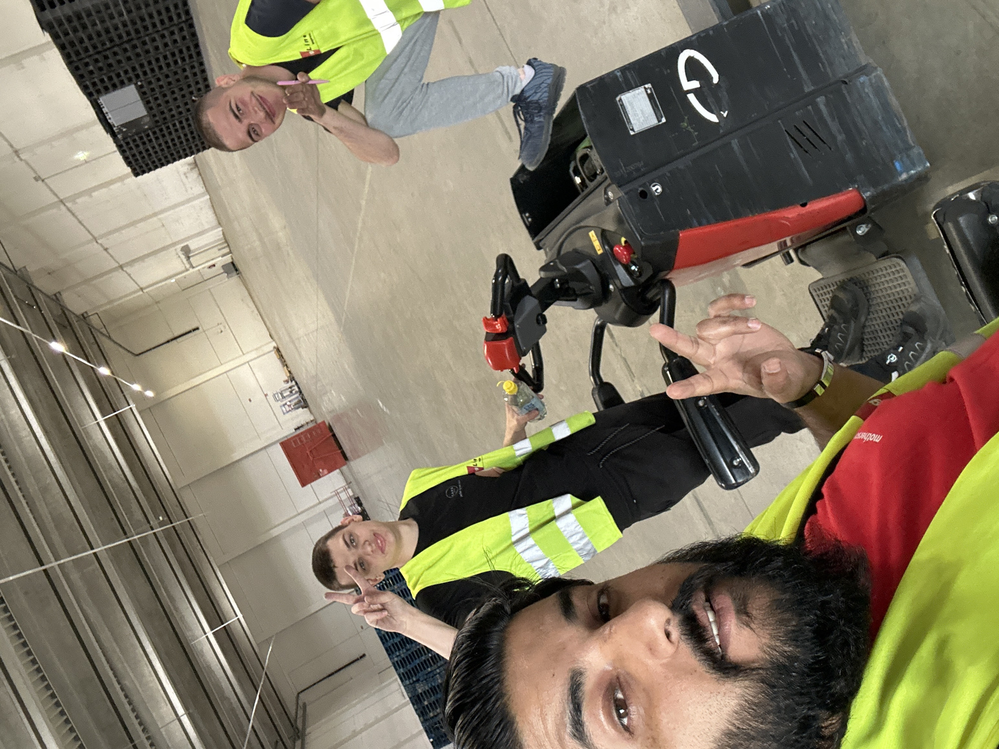 Three men are posing for a selfie in a large, empty warehouse, possibly a distribution center. The man in the foreground on the left is taking the photo, showing a portion of his face and wearing a bright yellow safety vest over a red t-shirt. He has a beard and is making a peace sign with his right hand. To his right, a second man is partially visible, also wearing a yellow safety vest over a black t-shirt and making a peace sign with his left hand. He is holding a clear plastic bottle with a yellow cap. Behind them, the third man is kneeling and smiling at the camera, wearing a yellow safety vest and dark pants. He is holding a pink object, possibly a pen. They are standing on a concrete floor. In the background, there are rows of stacked black pallets and some red storage containers. The ceiling of the warehouse has exposed beams and fluorescent lighting. The overall environment suggests an industrial or logistical setting. The man in the foreground has a black band on his left wrist. The warehouse appears to be an Amazon logistics center in Schweitenkirchen, Germany, given the branding visible on one of the pallet stacks.