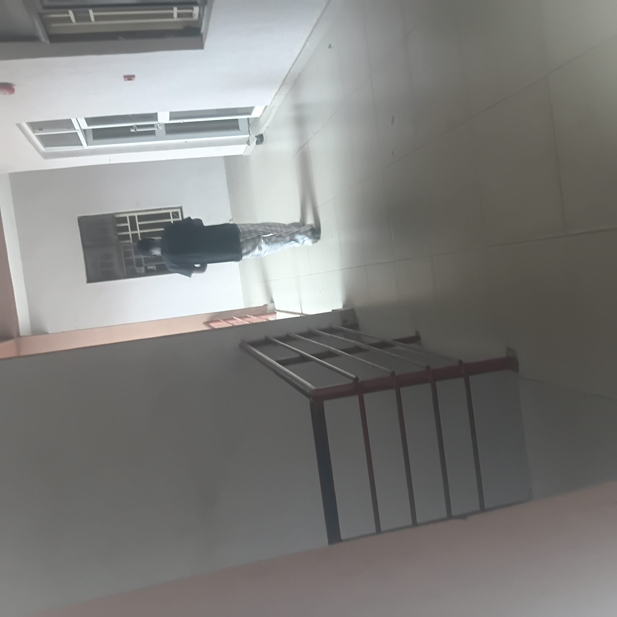 This media file captures an interior scene, likely a hallway or corridor within a building. The perspective is from a high angle, looking downwards. Dominating the frame is a person standing with their back to the camera, wearing a dark shirt and checkered pants. They appear to be looking downwards or at something on the floor.
The environment is constructed with light-colored walls and a tiled floor. There are windows visible on the walls, some with grilles, suggesting a functional rather than decorative purpose for these openings. A prominent feature in the lower right of the image is a set of railings, possibly belonging to a staircase or balcony, with reddish-brown supports and silver-colored horizontal bars. The lighting in the scene is somewhat dim, casting shadows and creating a slightly muted atmosphere.
There are no overt indicators of the time of day or weather conditions. The location context provided is Wukari, Nigeria, which might suggest a building typical of that region, though without more visual cues, specific architectural styles or cultural elements are not definitively discernible. The overall impression is one of a functional building interior with a single individual present. No visible text is present in the media.