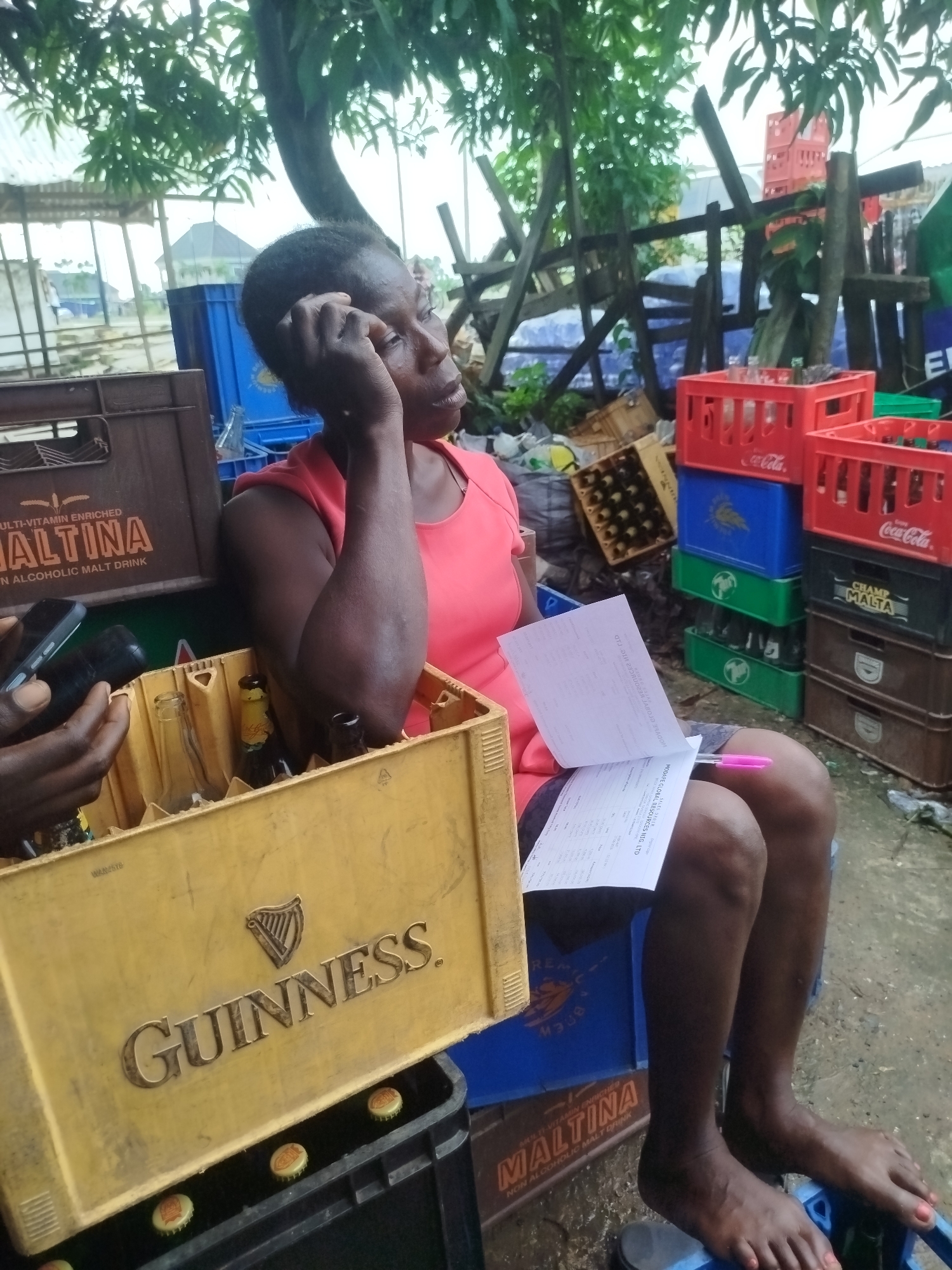 In Yenegoa, Nigeria, a woman sits amidst stacks of beer crates and bottles, seemingly overseeing operations. She is wearing a vibrant pink top and dark shorts, her posture relaxed as she holds a document and a pen. Her hand rests on her head, suggesting a moment of contemplation or perhaps fatigue. The scene is outdoors, under a leafy tree that provides some shade. The ground is wet, indicating recent rain.
Various crates and bottles are visible, including Maltina, Guinness, and Coca-Cola. Some bottles are empty, while others appear to be full or partially filled. A yellow Guinness crate is prominent in the foreground, containing several empty bottles. The presence of these beverages, along with the woman's engagement with a document, suggests a business transaction or inventory management is taking place. The background shows more crates, wooden structures, and some scattered debris, indicating a bustling but perhaps somewhat disorganized marketplace or distribution point. The overall impression is one of a hardworking individual managing a beverage distribution business in a tropical environment. The lighting suggests it might be daytime, possibly after a rain shower.
