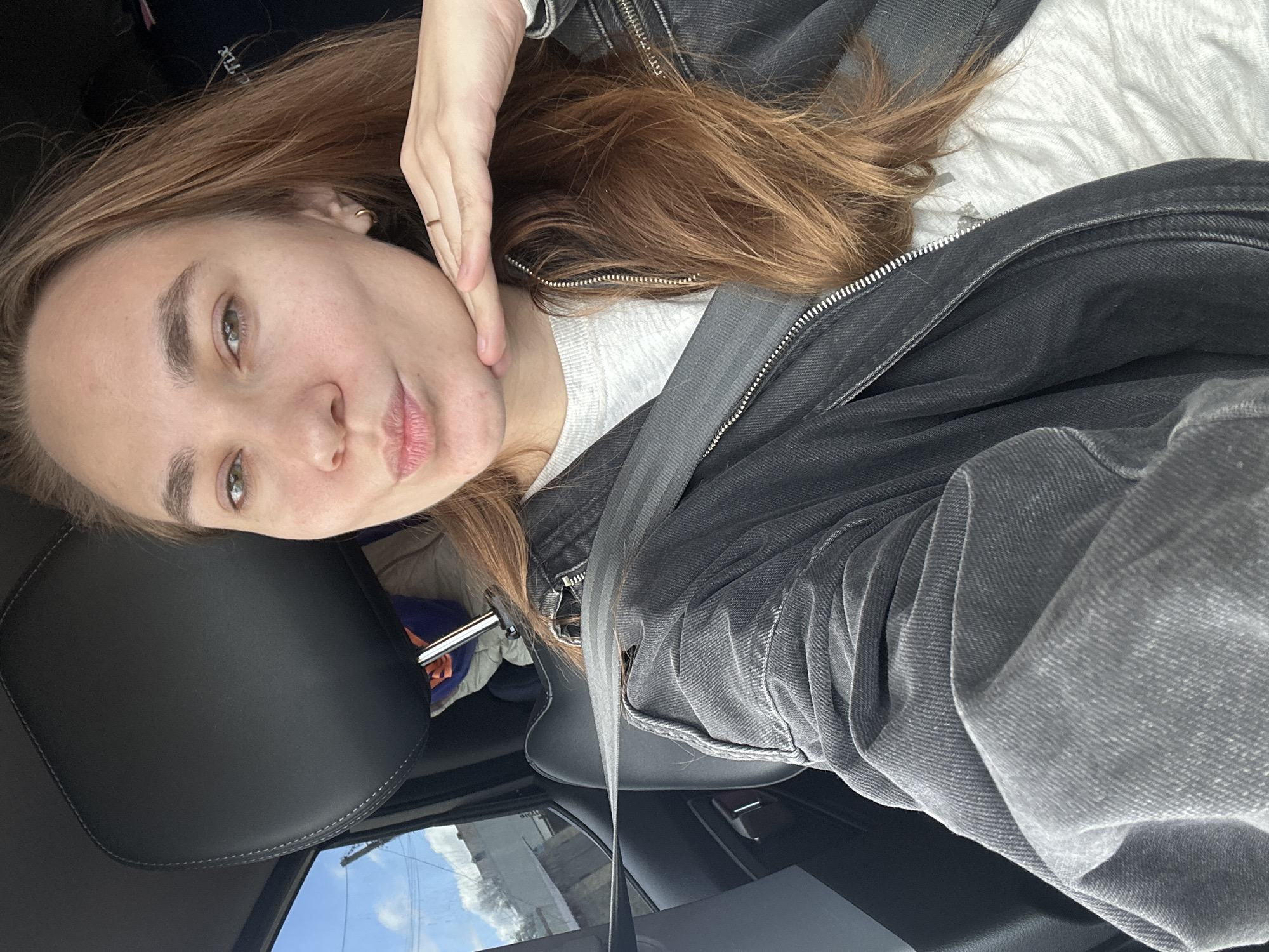 A woman with shoulder-length brown hair, wearing a light gray t-shirt and a black denim jacket, is in a car. She is wearing a seatbelt and appears to be taking a selfie. The scene shows the car's interior, including the headrest, door, and a glimpse of the sky outside the window. There is no indication of the location being in Chaikovsky, Russia.