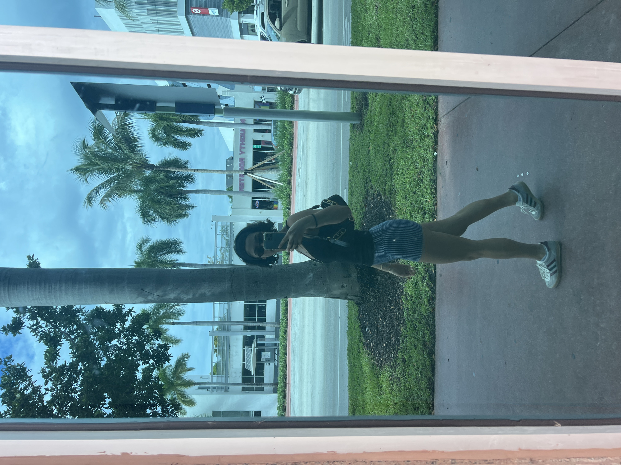 A person stands on a sidewalk in front of a grassy area in Miami Beach, United States of America, reflected in a window. The person is wearing a dark top and denim shorts with Adidas sneakers. They are holding a cell phone, presumably taking the photograph. Behind the grass and sidewalk are buildings, palm trees, and a street. A Target store sign is visible in the background, in addition to other commercial buildings.