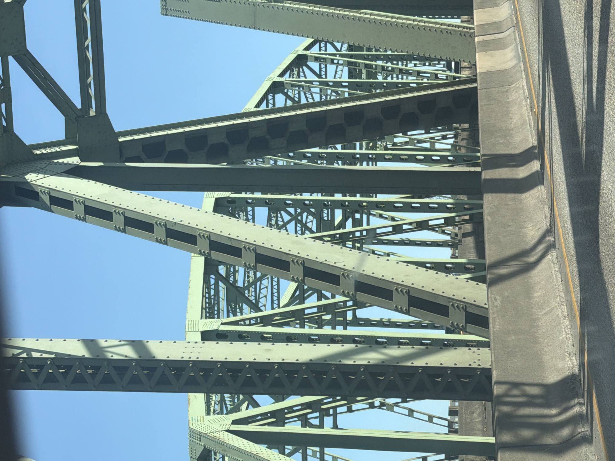 This is a close-up, angled photograph of the steel structure of a bridge, likely taken from a vehicle moving across it. The dominant subjects are the interconnected girders and beams of the bridge, painted a muted green. The sky is a clear, bright blue, suggesting a sunny day. The time of day appears to be daytime due to the clear sky and the shadows cast on the bridge structure. The scene is urban or semi-urban, given the presence of a large bridge and what appears to be a roadway on the right side of the frame, marked with a yellow line and concrete barriers. There are no people visible. The overall impression is one of industrial strength and engineering. The location is suggested to be Vancouver, USA. There is no visible text in the image.