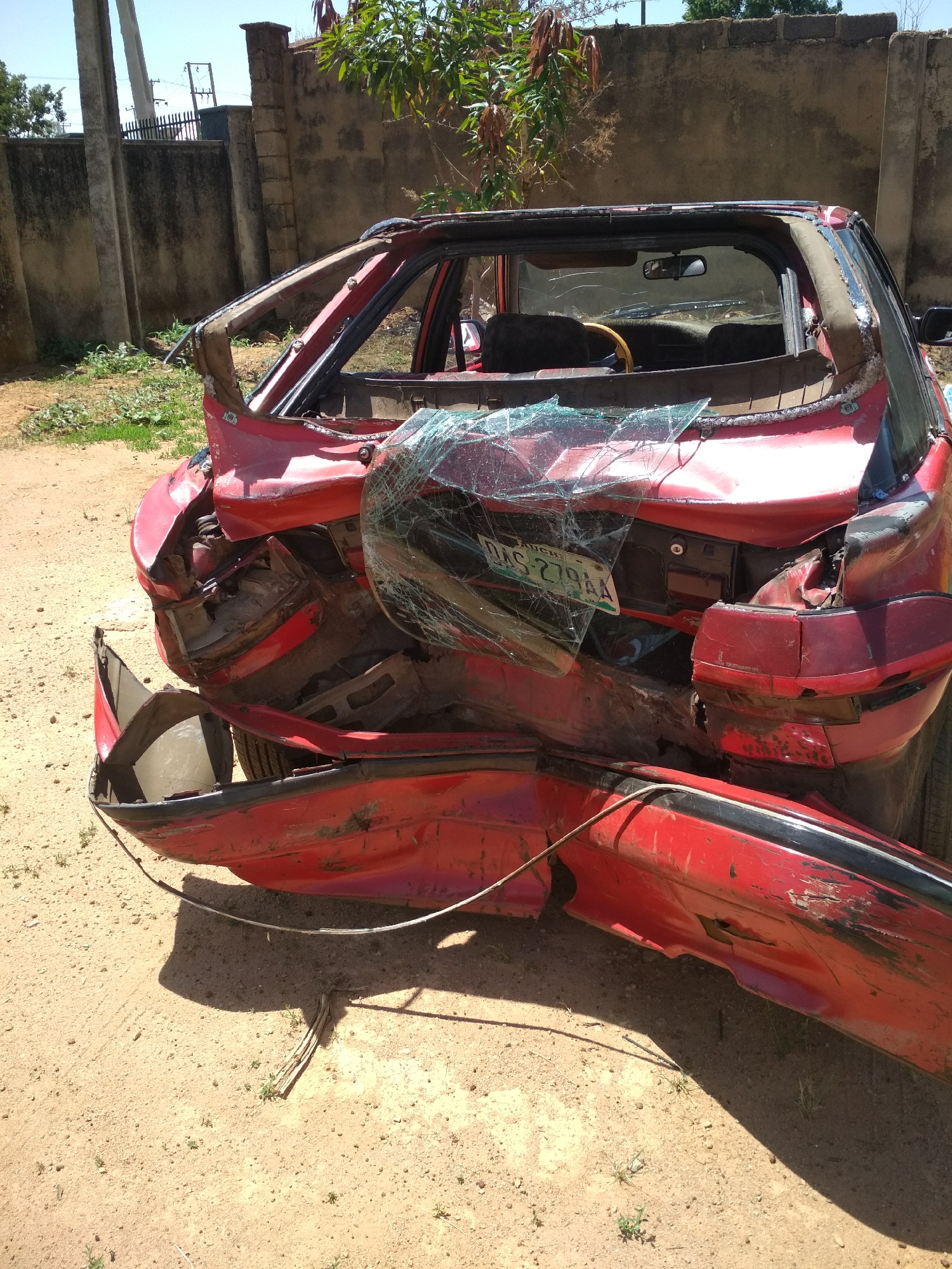 The image captures a severely damaged red car, likely a sedan or hatchback, stationary outdoors under bright daylight. The vehicle has sustained catastrophic damage to its rear and roof sections. The entire rear end is crushed, twisted, and torn open, with the rear bumper detached and lying bent on the ground next to the car. Shattered glass from the rear window is scattered over the trunk and chassis, while fragments of glass are also visible around the interior. The roof pillars are visibly bent and broken, suggesting a rollover or severe impact.
Through the mangled roof and windows, a wooden-rimmed steering wheel and dark-colored seats are discernible inside the vehicle. A Nigerian license plate is partially visible on the crushed trunk, displaying the text "BAUCHI" at the top and "DAG-279AA" below it.
The car rests on dry, sandy ground, possibly a dirt road or an unpaved area. In the background, a high, unpainted concrete wall spans horizontally, with green foliage and trees visible behind and above it. To the left, several tall concrete utility poles stand against a clear, bright blue sky. The harsh shadows cast on the ground indicate a sunny, clear daytime, likely mid-day. The scene depicts the aftermath of a major vehicle accident.