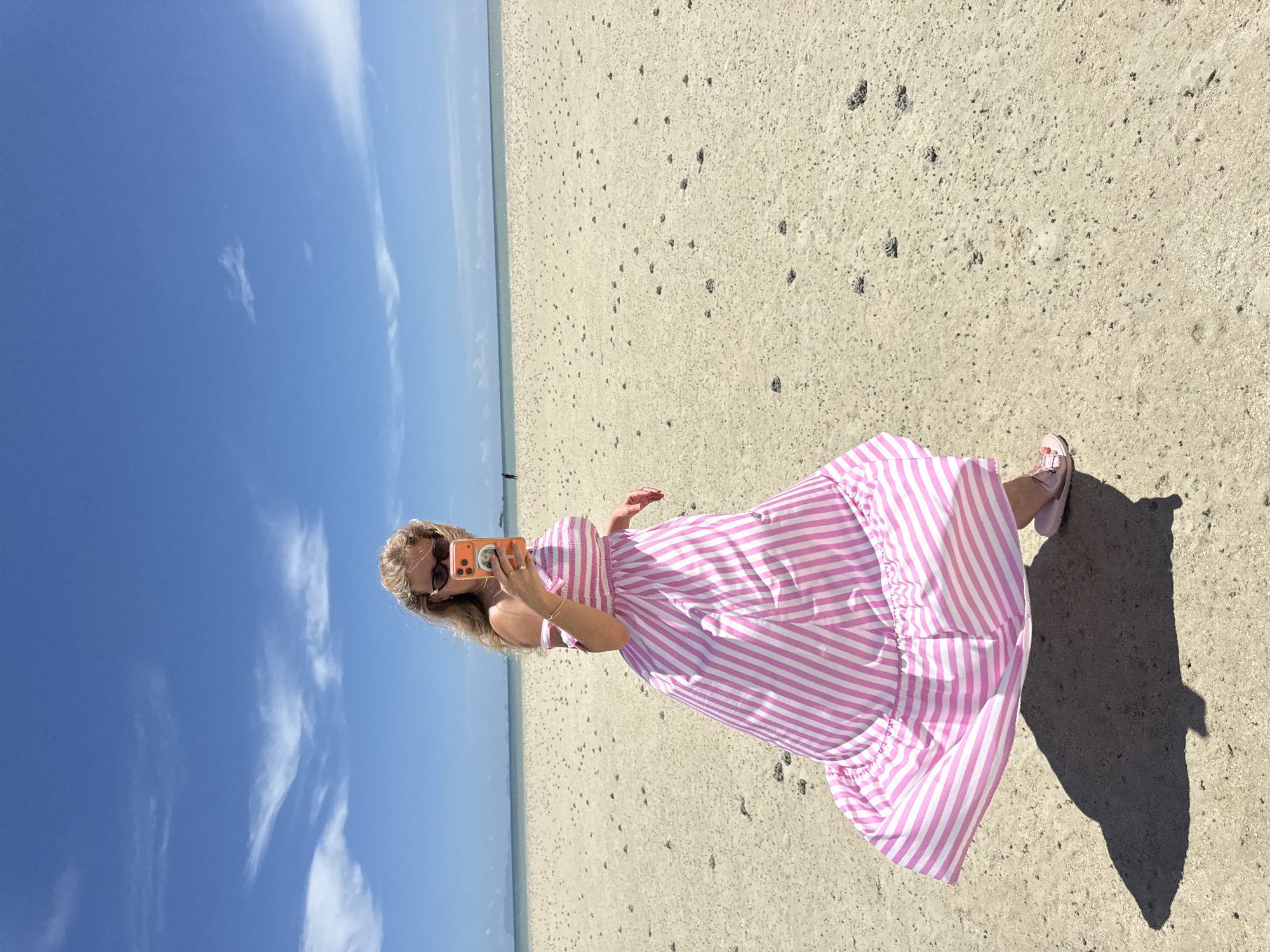 A woman with blonde hair is taking a selfie on a sandy beach under a bright blue sky with sparse clouds. She is wearing a pink and white striped maxi dress, sunglasses, and pink sandals. Her phone, with an orange case, is held up to take the photo. The beach is largely empty, with some small dark objects scattered across the sand. In the distance, the ocean meets the horizon, and faint outlines of land are visible. The overall setting appears to be a tropical or sub-tropical coastal area.