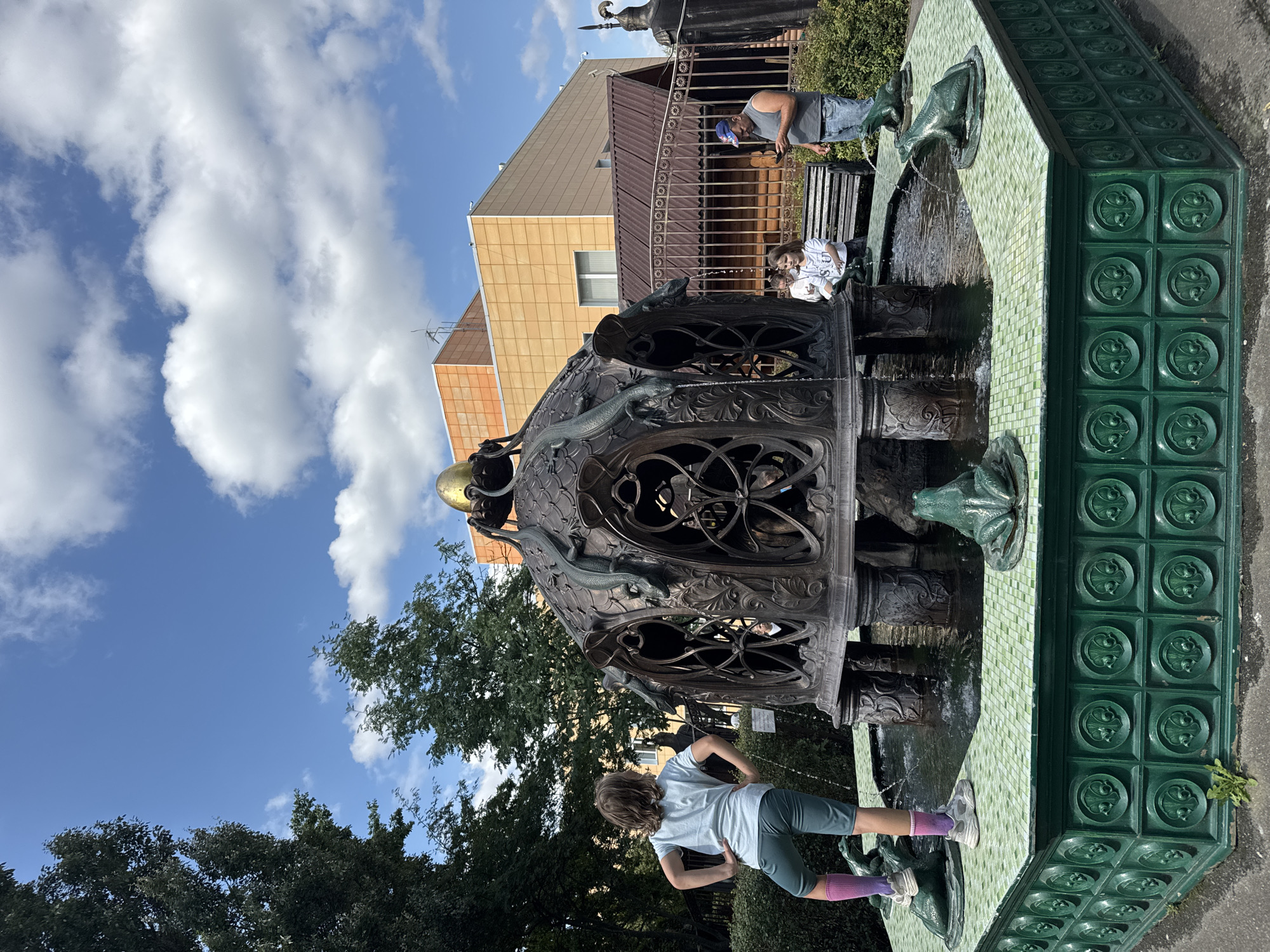 The image shows a decorative fountain with a young girl standing on its edge and two women and a man nearby. The fountain features a large bronze structure with intricate metalwork, topped with a golden orb. Several bronze lizards are depicted climbing the structure, and water spouts from their mouths. The base of the fountain is lined with green tiles, and there are frog sculptures around the edge. The background shows a building with an orange facade, trees, and a clear blue sky with white clouds. The scene is set in a public outdoor space, likely a park or square, in the city of Sovkhoz imeni Lenina, Russia. The young girl is facing away from the camera, with her hands on her hips. One woman is sitting on a bench, and the other is standing beside her, looking towards the fountain. The man is standing further away, looking at his phone.