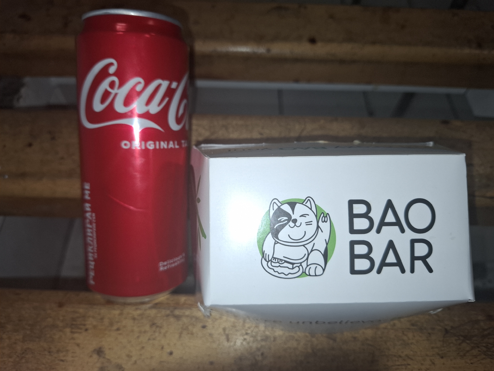 A Coca-Cola can and a white box from "Bao Bar," a restaurant in Sofia, Bulgaria, are sitting on a wooden bench. The can is red with white lettering. The box is white with a cartoon of a cat holding a bao in a green circle and the words "BAO BAR" in black lettering. The background is blurred, but appears to be a wooden structure.