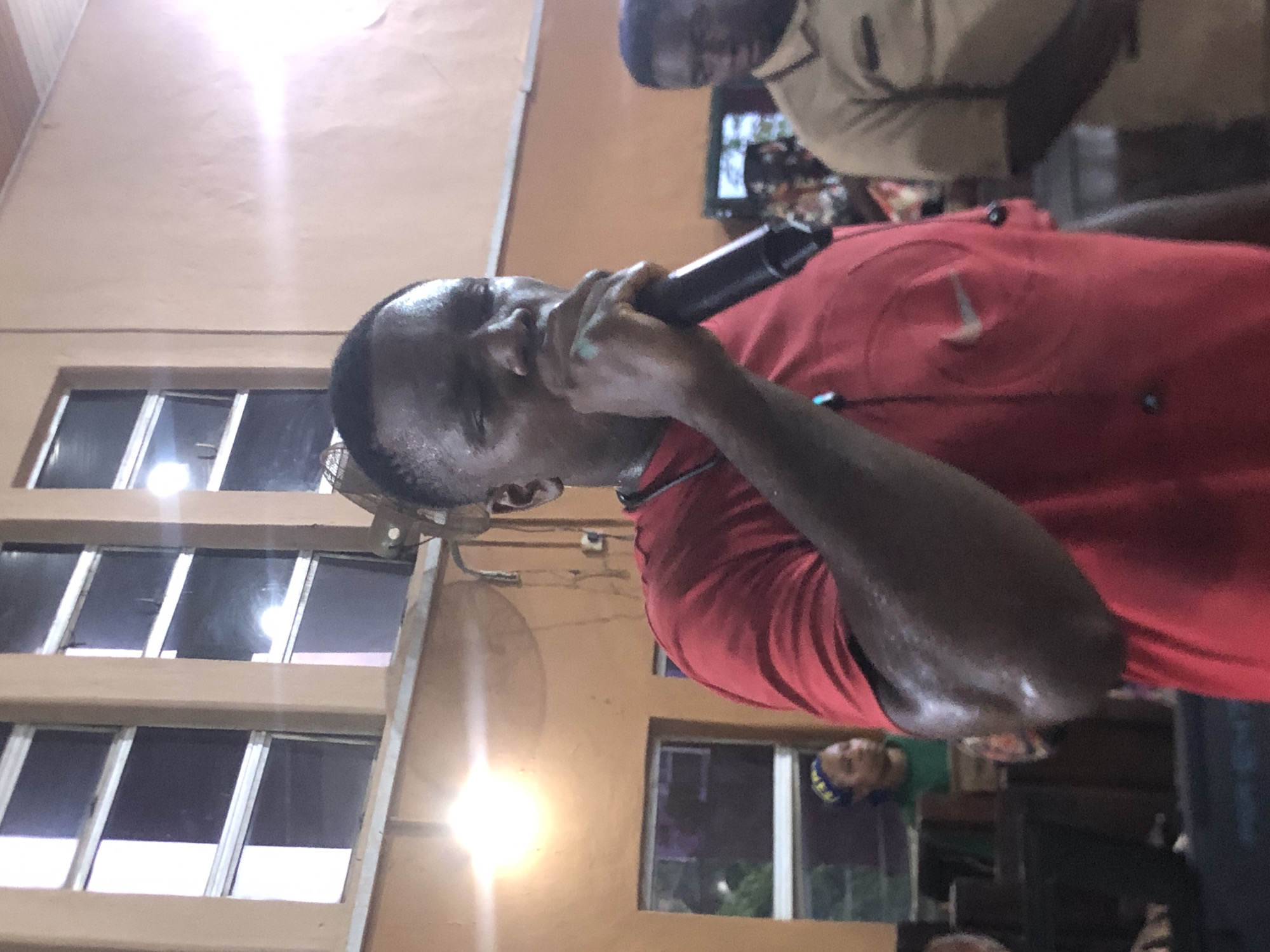 A man is singing into a microphone in Nsugbe, Nigeria. He is wearing a red shirt and standing indoors near a wall with windows. Other people are present in the background, including one person wearing a green shirt and a blue and yellow head covering. A ceiling fan is visible on the wall.