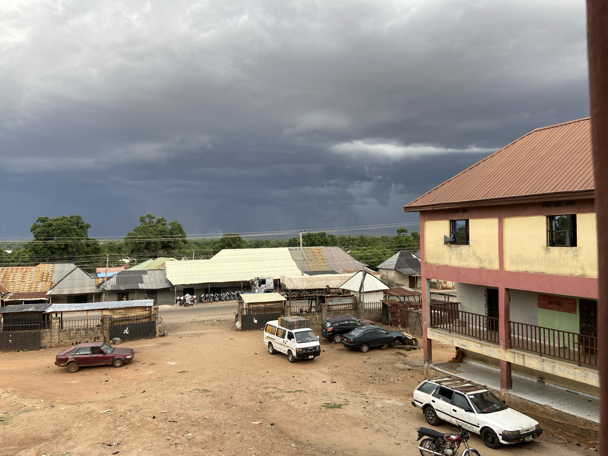 The image shows a street scene in Mararaba, Nigeria, under a cloudy sky with dark storm clouds. The ground is mostly dirt. A maroon car is parked in the dirt area on the left side of the frame. In the center, there is a white van with luggage tied to the roof and two dark colored cars parked nearby. Further in the background, there are people walking on the side of a road next to parked motorcycles. A two-story building with balconies is situated on the right side of the frame. In front of the building, a white Toyota station wagon and a motorcycle are parked. A person is sitting on the ground under the building's balcony. Buildings with corrugated metal roofs are across the road.