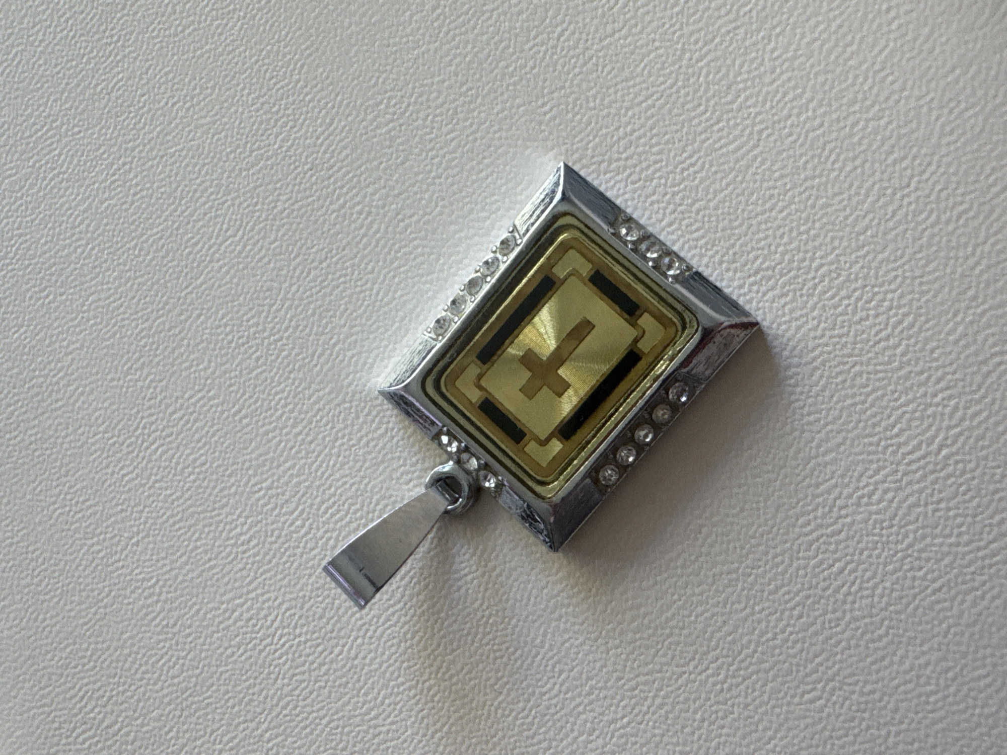 The image shows a close-up, top-down view of a pendant lying on a textured, light-colored surface, likely a table or cloth. The pendant is diamond-shaped with a silver-colored metallic frame. The frame is adorned with small, clear stones, possibly diamonds or rhinestones, along its edges. Inside the frame, there is a rectangular design with a gold-colored centerpiece featuring a cross-like symbol in a darker gold hue. Two black rectangular accents are placed on either side of the gold centerpiece within the frame. A silver-colored bail or loop is attached to the bottom point of the pendant, allowing it to be hung. The overall impression is of a piece of jewelry. There is no visible text. The lighting suggests an indoor setting, possibly during the daytime due to the bright illumination. The setting is not explicitly identifiable beyond the pendant and its immediate surface.