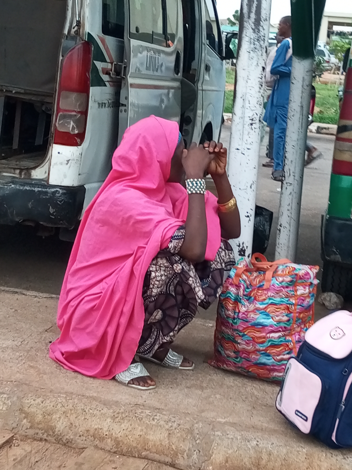 A woman wearing a bright pink headscarf and patterned skirt is crouched on a concrete surface, next to a large, colorful bag and a dark blue backpack with pink trim. Her hands are clasped in front of her face, and she is wearing beaded bracelets on her left wrist and a gold bangle on her right. A white van with a red taillight and green and white decals is parked behind her. A man in a blue traditional outfit stands near a pole in the background, and other people are visible further away. The environment appears to be an outdoor area, possibly a transportation hub or market, in Gombe, Nigeria.