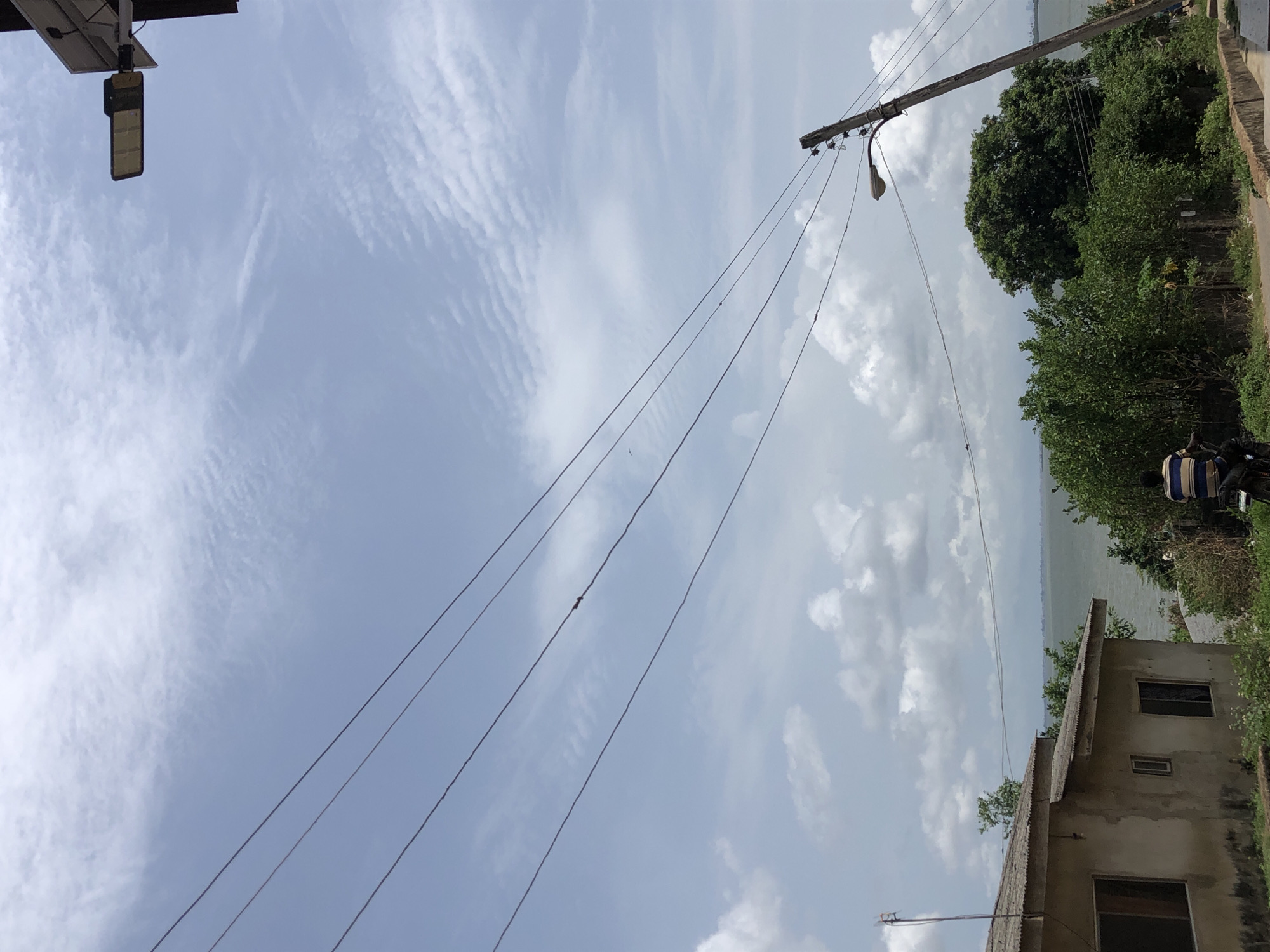 The scene depicts a view of a partially cloudy sky, power lines stretching across the frame, and a glimpse of what appears to be a body of water in the distance. A wooden utility pole with a streetlight attached stands at an angle. Vegetation, including trees and bushes, is visible to the right side of the frame. A portion of a building with a dilapidated roof and visible windows is also seen on the right. In the foreground, the top of a solar panel and a dark-colored street lamp are present. A person on a motorbike is visible next to the bushes. The location is Unknown, Unknown.