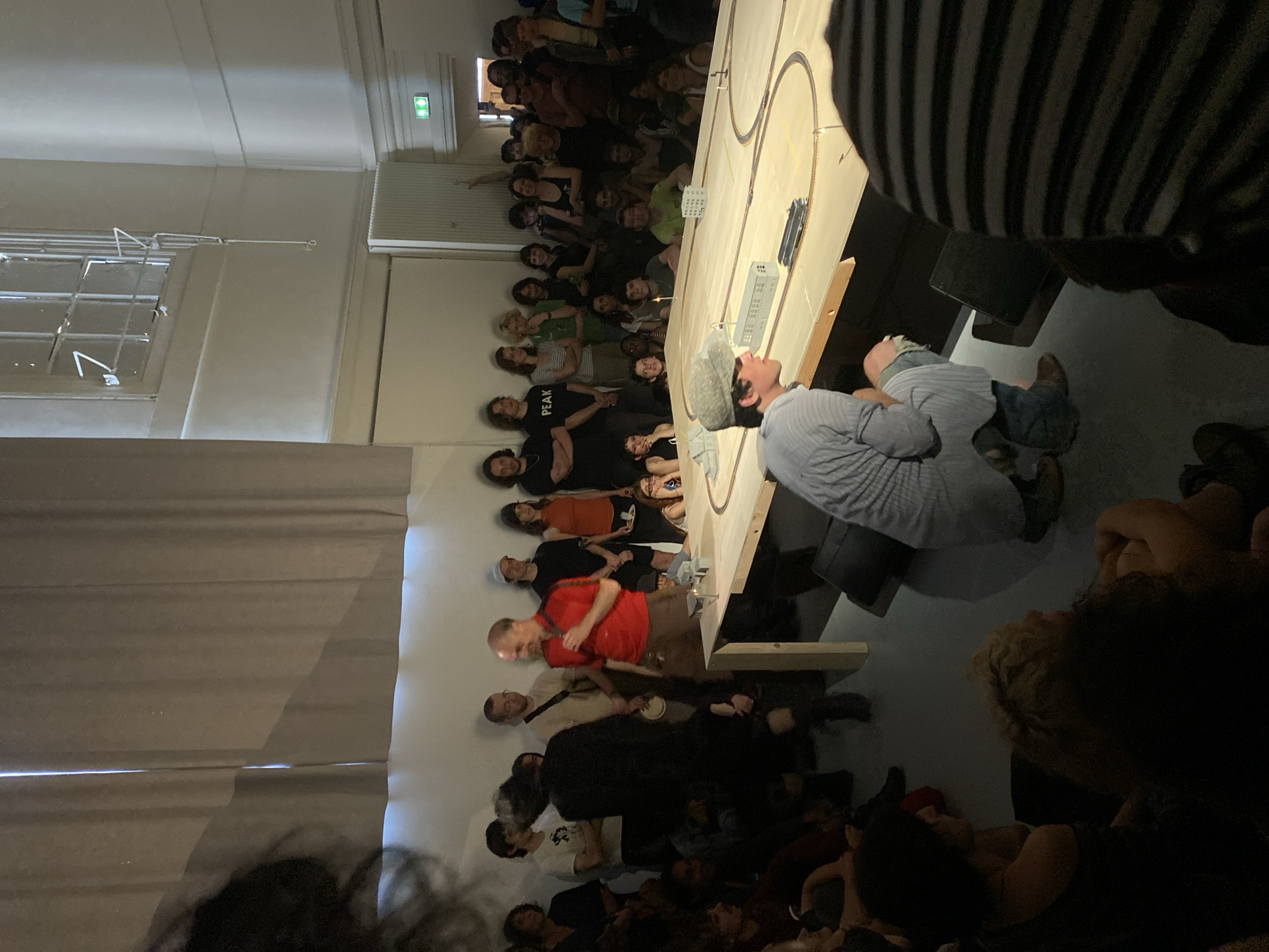 The photograph depicts a crowded indoor scene. A man wearing a grey shirt and a hat sits at a long table where a model train set is set up, and the crowd is watching the man and the train. Many people are standing or sitting on the floor. Some people are wearing black t-shirts with white writing on them. The environment is a large room with a window on the left side and an exit sign is visible. The image was taken inside the Musée des Arts et Métiers in Paris, France.