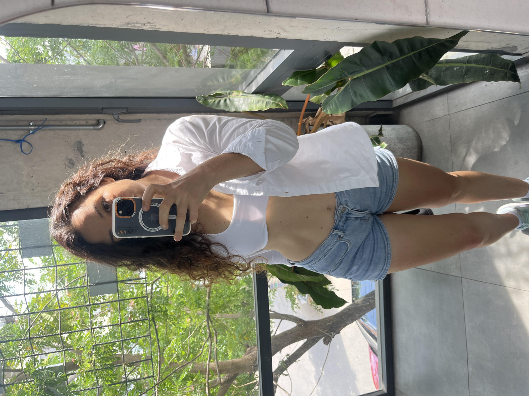 A young woman with curly brown hair takes a mirror selfie in what appears to be a modern apartment with large windows overlooking greenery. She is wearing a white crop top, an unbuttoned white shirt, and denim shorts. The background shows trees and foliage, suggesting an outdoor urban environment. The image is well-lit with natural light. The specific location of Phu Thuy Ward, Vietnam, cannot be determined from the visual information provided in the image.