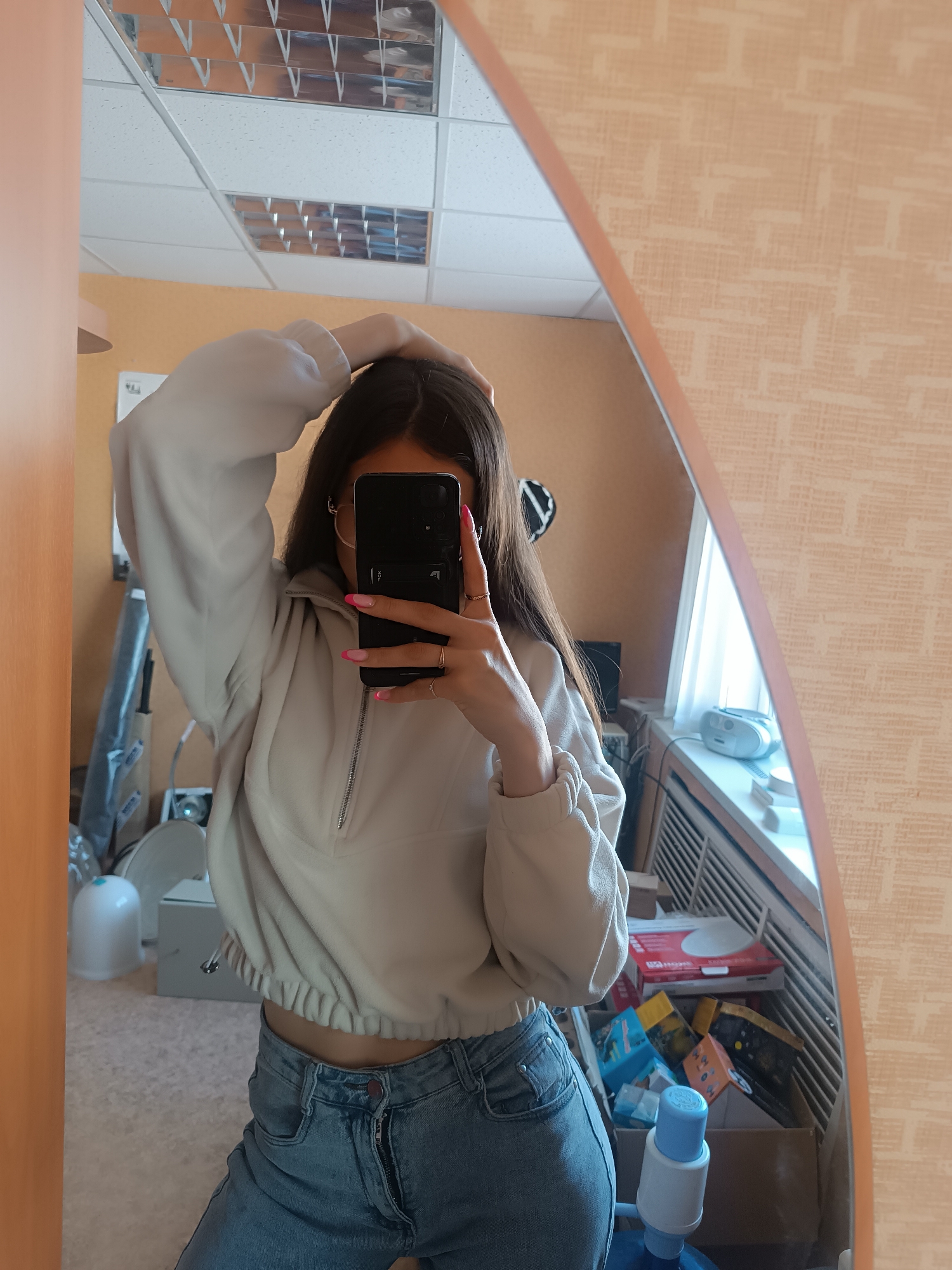 A young woman is taking a selfie in a mirror. She is wearing a beige, cropped sweatshirt with a zipper and light blue jeans. Her left arm is raised and her hand is touching her head, while her right hand is holding a smartphone in front of her face, obscuring it from view. She has long, dark hair and wears glasses. Her fingernails are painted with a pink French manicure.
The mirror has an oval shape with an irregular frame. The background is a room with light-colored wallpaper, a white ceiling with fluorescent lights, and a window with a radiator underneath. There are various objects scattered around the room, including boxes, a water dispenser, and a vacuum cleaner.
The location is an indoor setting, possibly a home or office. The city is Unknown, Unknown.