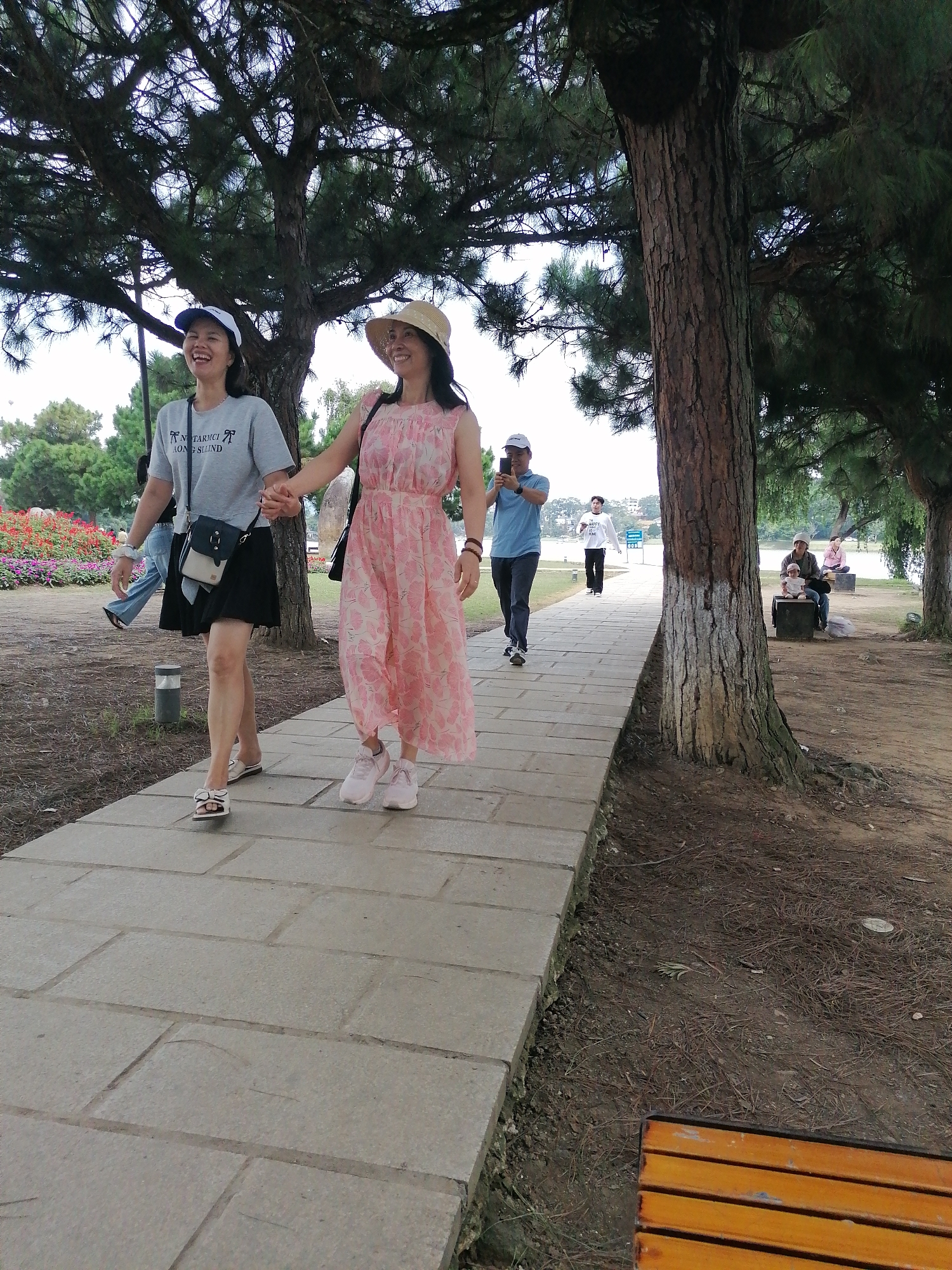 Two women walk hand-in-hand on a stone pathway in a park near Xuan Huong Lake in Da Lat, Vietnam. The woman on the left wears a grey t-shirt, a black skirt, and a blue and white cap. The woman on the right wears a pink floral dress and a straw hat. Both are smiling and appear to be enjoying themselves. In the background, several other people are visible, including a man taking a photo with his phone, and a group sitting on a bench near the lake. The park is shaded by large pine trees, and the ground is covered with pine needles. Flowers are visible on the left side of the pathway.