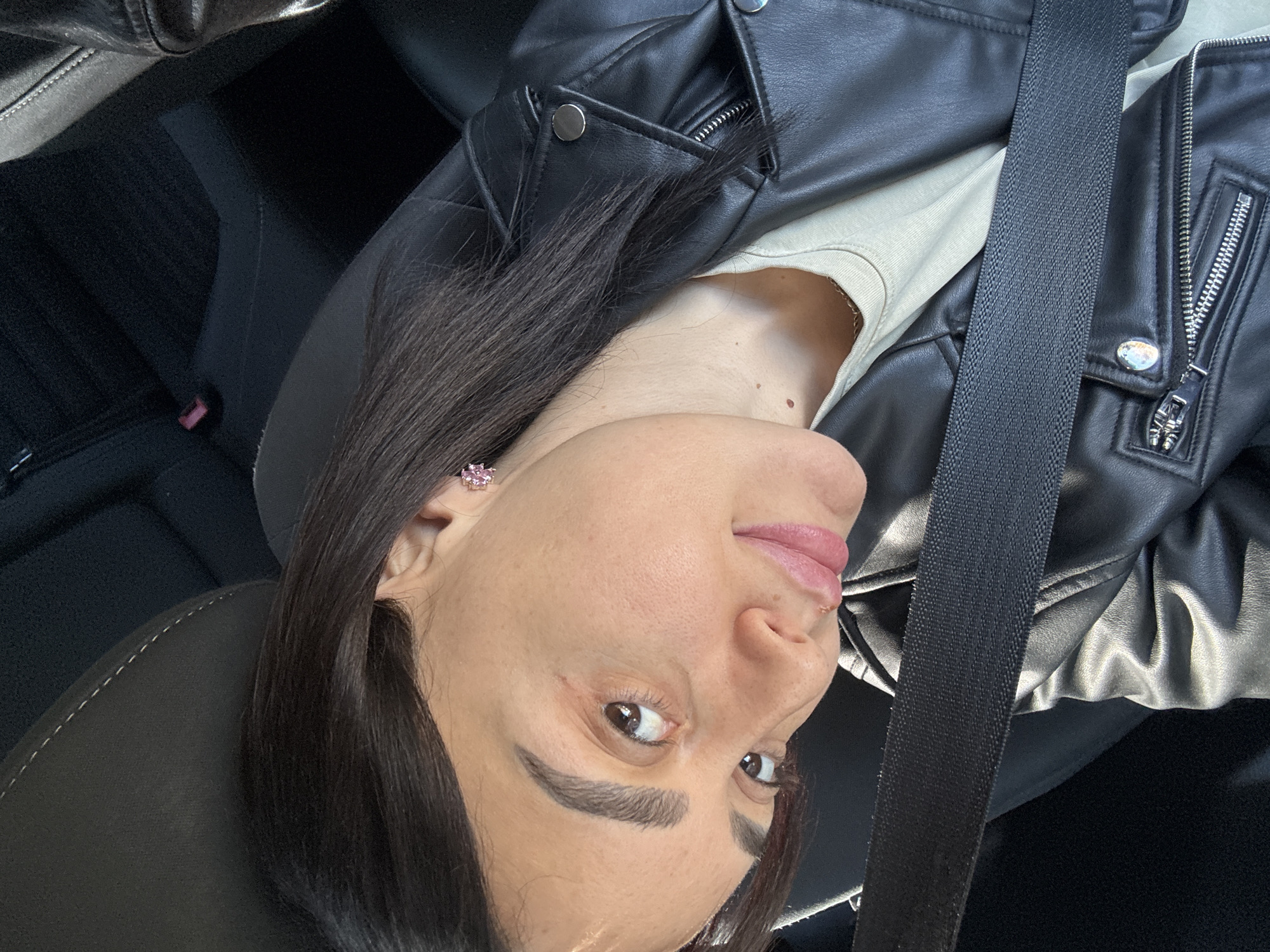 A woman with dark hair and fair skin is shown in a close-up selfie, taken from a low angle. She is wearing a black leather jacket and a light-colored top, with a seatbelt diagonally across her torso. A small pink earring is visible in her left ear. She is seated in a car, as indicated by the seatbelt and the car seat visible in the background. The location is not discernible from the image.