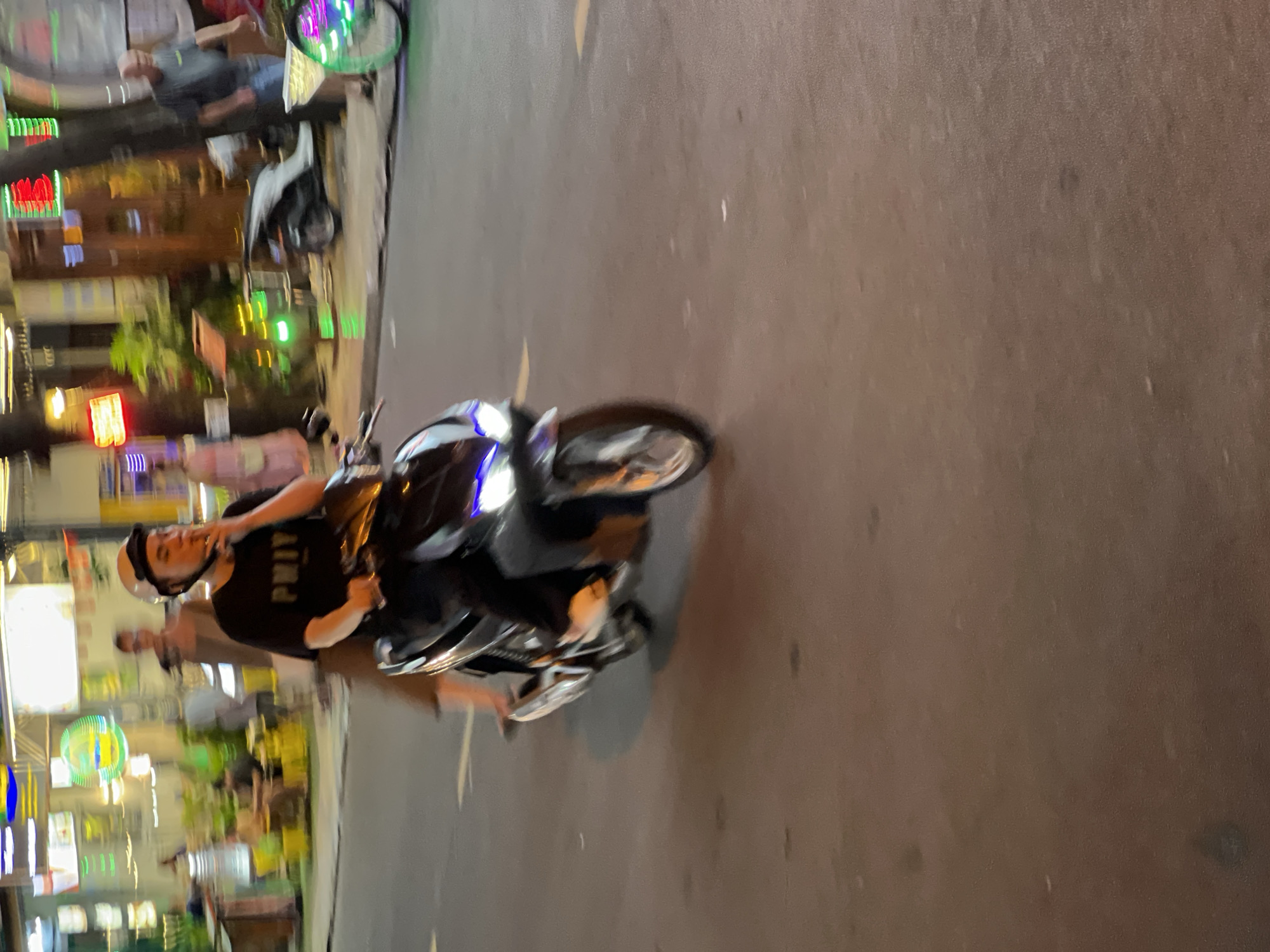 A person is riding a dark-colored scooter with blue LED headlights on a street at night. The person is wearing a black t-shirt with white lettering that appears to read "PMIY" and a white helmet. They are holding a cigarette to their mouth. The scooter is in motion, indicated by the blur. In the background, there are neon signs and people walking on the sidewalk. The street is paved and has yellow lane markings. The lighting suggests an urban commercial area, likely a busy street in Nha Trang, Vietnam, known for its vibrant nightlife and street food scene. The overall atmosphere is lively and typical of a Southeast Asian city at night.