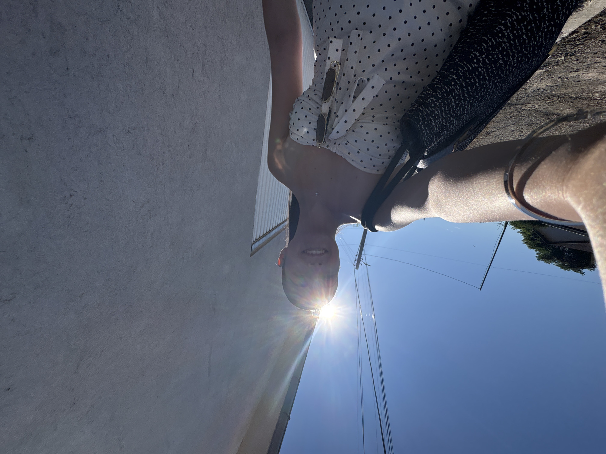 A person is taking a selfie in the city of Bulgarovo, Bulgaria. The person is wearing a white polka dot dress and holding a black bag. They are standing outside on a sunny day, with a bright sunburst in the background. The person is smiling and their face is partially obscured by the sun. The environment includes a concrete structure overhead, power lines, and a clear blue sky. The image is taken from a low angle, looking upwards.