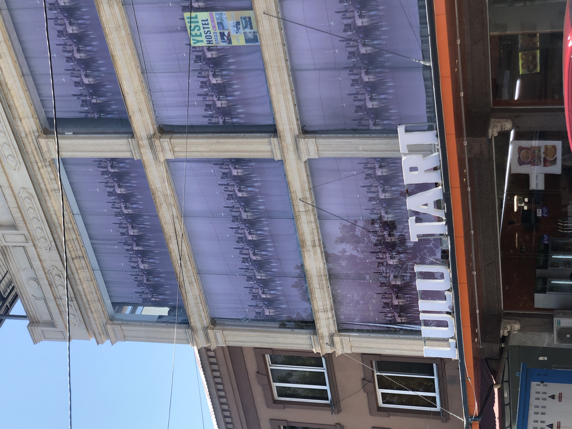 The image captures the exterior of a building in Tashkent, Uzbekistan, featuring a prominent sign reading "LULU TART". The building's facade is adorned with large, purple-toned panoramic images of a cityscape with reflections. To the right, a green sign for "YESIL HOSTEL" is visible. The building is multi-storied, with several floors of windows and decorative architectural elements. A chain hangs from the building, possibly for support or decoration. The lower part of the image shows the entrance to what appears to be a shop or restaurant, with glass doors and displays. Electrical boxes and air conditioning units are also present. The overall scene suggests a commercial area within the city.