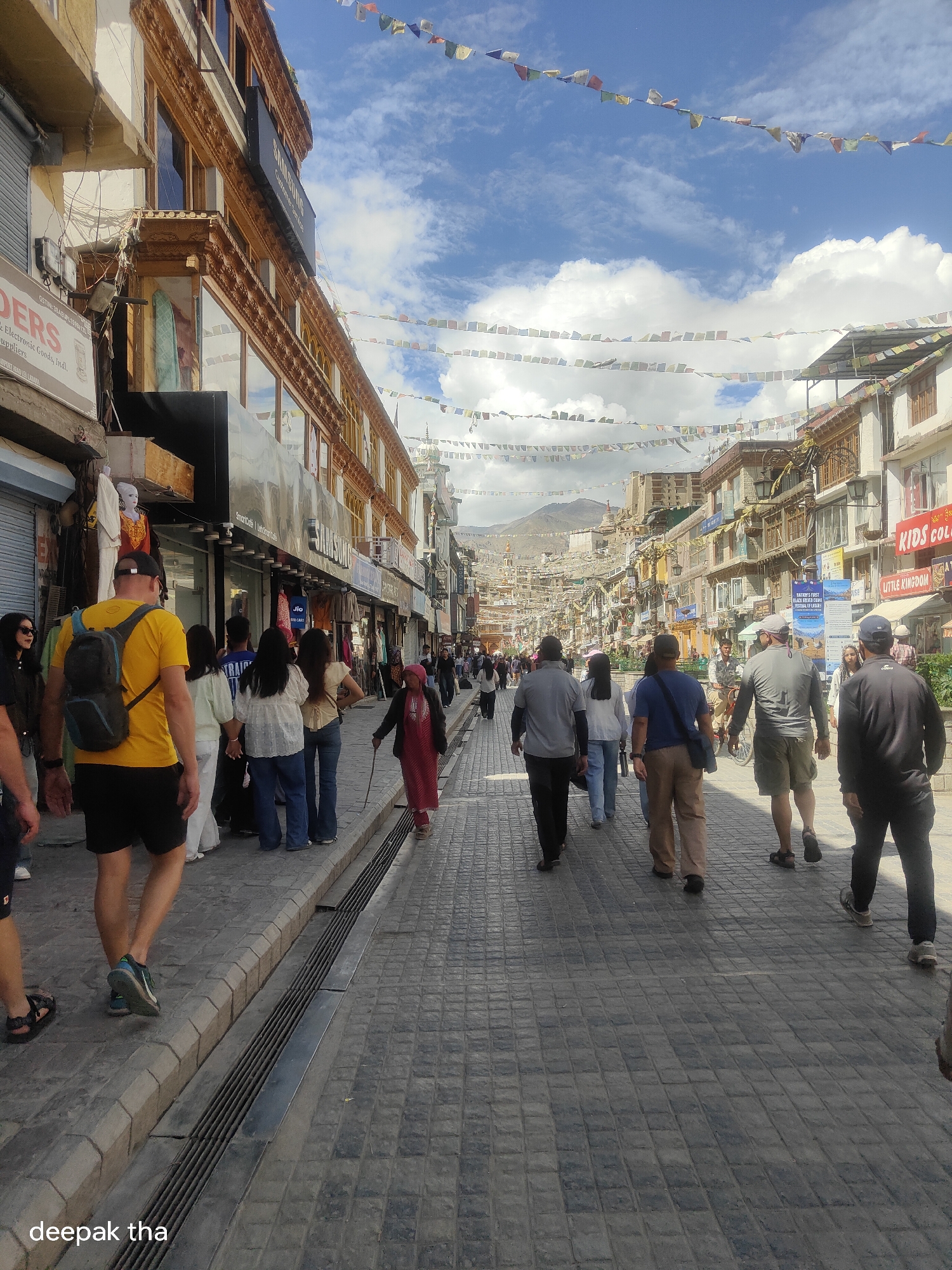 The image depicts a bustling street scene in the city of Leh, India, likely on the main market road. The street is paved with cobblestones and lined with multi-story buildings housing shops and businesses. Prayer flags are strung across the street overhead, adding a vibrant splash of color against the blue sky dotted with white clouds. Numerous people are walking along the street, some singly, some in small groups, suggesting a lively atmosphere. The architecture of the buildings appears to be a mix of traditional Ladakhi style and more modern commercial construction. The overall impression is one of a vibrant, active urban environment within a mountainous region.