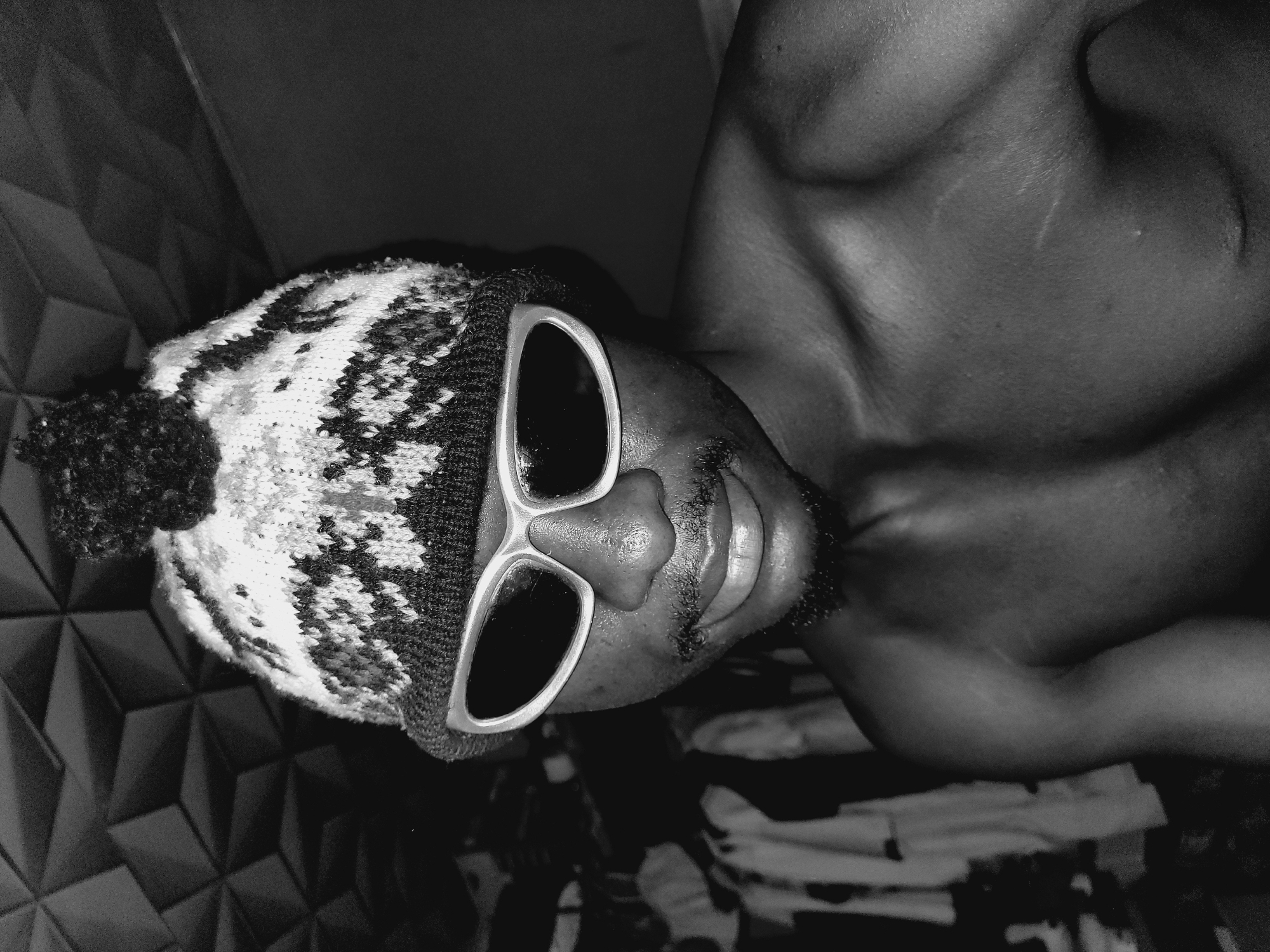A monochrome, angled close-up captures a shirtless person with dark skin, wearing a patterned knit hat with a pom-pom and oversized, light-colored sunglasses. The person is facing upward, with part of their chest and neck visible. The background includes a textured wall and possibly a patterned surface on which the person is lying. The location is not discernible from the image alone, and the city of Bukuru, Nigeria, cannot be confirmed.
