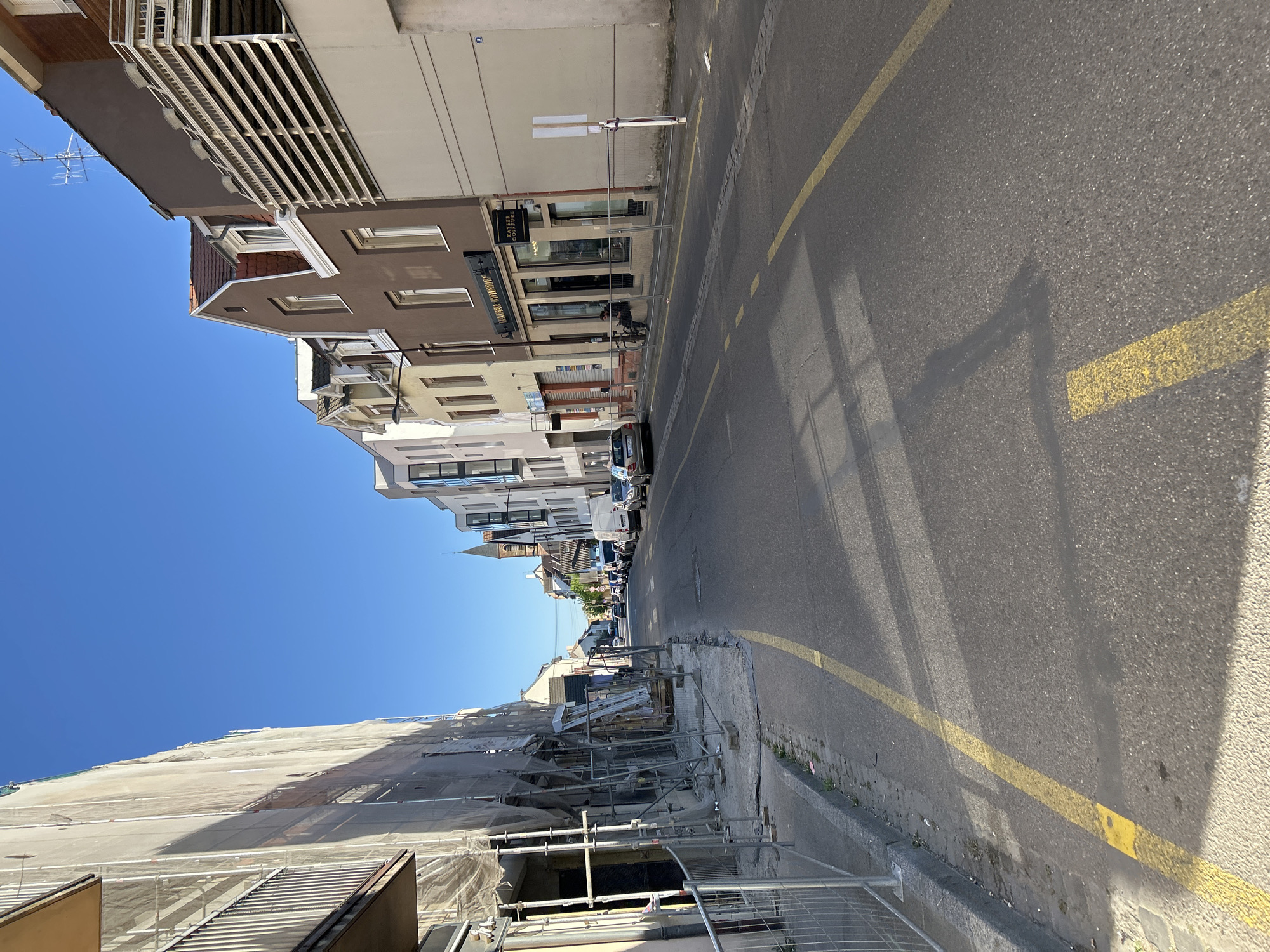 A vibrant, sunlit urban street scene unfolds under a clear blue sky. The image captures a paved road extending into the distance, bordered by multi-story buildings on both sides.
On the left, a large building is extensively covered with white construction scaffolding and protective netting, indicating ongoing renovation or construction. Various building materials and equipment are visible at ground level.
The right side of the street features a row of multi-story buildings with varied architectural styles. A prominent brown building closer to the foreground showcases a distinct balcony or awning structure and multiple windows. Further down, another brown building houses ground-floor businesses. Visible text on storefront signs includes "MAISON ALONSO BOUTIQUE" and "PRODUITS BASQUES." Another sign appears to read "COSMETIQUE" or "COIFFURE," and a black sign bears the text "SARL ANTHONY G...". Several antennas are visible against the sky above these structures.
The asphalt road is clearly marked with bright yellow painted lines, delineating lanes or parking areas. Strong, defined shadows of buildings and unseen elements stretch across the pavement, suggesting it is a sunny daytime, likely morning or late afternoon. A few cars are parked along the right curb further along the street. In the background, a church steeple rises above the rooftops, hinting at the historical character of the area.