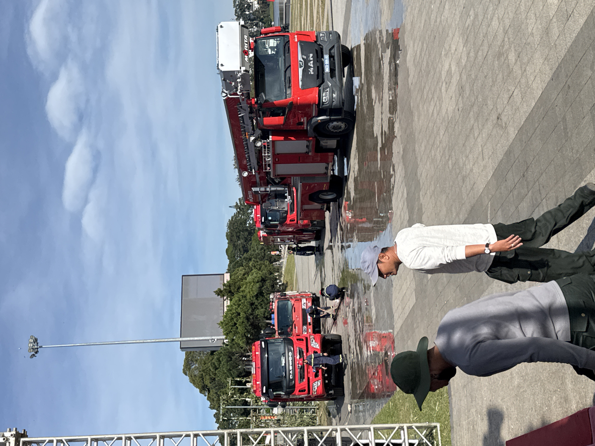 The image depicts an outdoor scene in Da Lat, Vietnam, likely a public space or event area, characterized by a paved ground surface with numerous puddles of water. Several red fire trucks are present, some with their ladders extended. Three individuals in firefighter uniforms are visible near the trucks, with one appearing to be cleaning or attending to the ground. Two additional men, dressed in civilian attire, are in the foreground, one wearing a dark green hat and a gray long-sleeved shirt, the other in a light gray long-sleeved shirt and dark pants. In the background, a large screen is mounted on a structure, and a tall lamppost stands prominently. Trees and a body of water are also visible in the distance, suggesting a park or lakeside location within the city. The sky is partly cloudy.