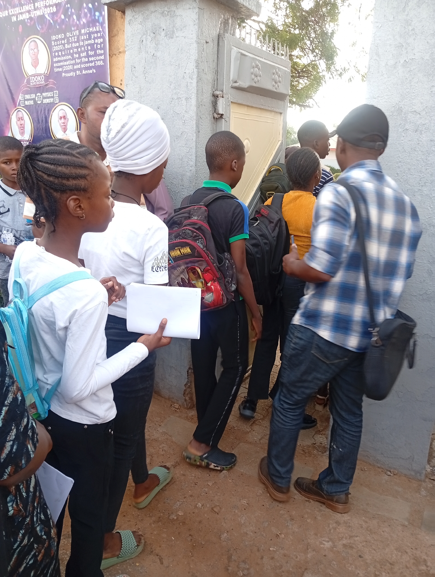 A group of young people stand outside a gray building in an area of reddish dirt in the city of Unknown, Unknown. Some are wearing backpacks, and at least one is holding a piece of white paper. At least one adult is present. A banner on the wall highlights the academic performance of someone named "Idoko Olive Michael" in JAMB-UTME 2026, noting scores in English, Physics, Math, and Chemistry. A gate is partially open.