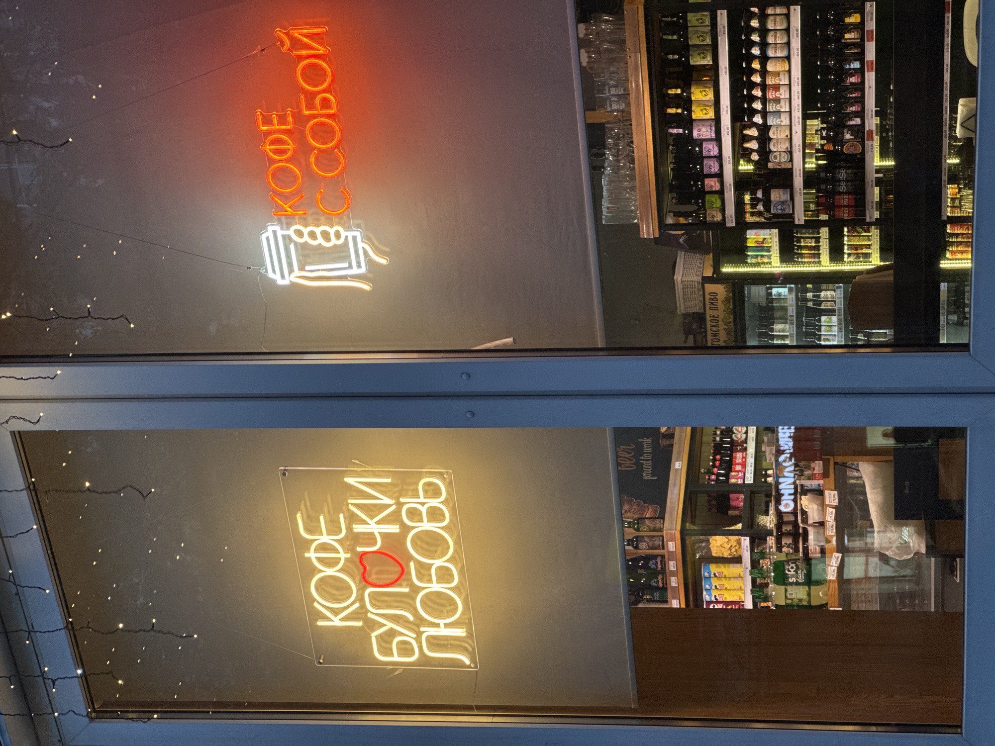 The image captures a nighttime view looking through a shop window, revealing an interior well-lit by artificial light and neon signs. The overall scene suggests a cafe or a small shop.
On the left side of the window, a bright white neon sign displays "КОФЕ БУЛОЧКИ ЛЮБОВЬ" (Coffee Buns Love) with a red heart icon integrated into the word "БУЛОЧКИ" (buns). To the right, another neon sign, orange and white, reads "КОФЕ С СОБОЙ" (Coffee To Go) and features a graphic of a coffee cup with steam. Both signs are attached to the interior side of the glass.
Behind the window, the interior of the establishment is visible, primarily showcasing shelves filled with various bottled beverages, likely beer, given the visible text and labels. Some bottles have price tags attached. Stacked drinking glasses can be seen on a shelf near the top right. Another sign inside reads "beer forced to work" and "ГОМСКОЕ ПИВО" (Gomskoe Beer). Various snacks and other products are also displayed on shelves and in refrigerated units.
Fairy lights are strung up on the exterior, possibly around the window frame, reflecting dimly on the glass surface. The setting appears to be urban, likely a street-facing shop, with no people clearly visible. The strong illumination from the neon signs and interior lights suggests it is either evening or night. The image is taken at a slight upward angle, showing parts of the window frame.