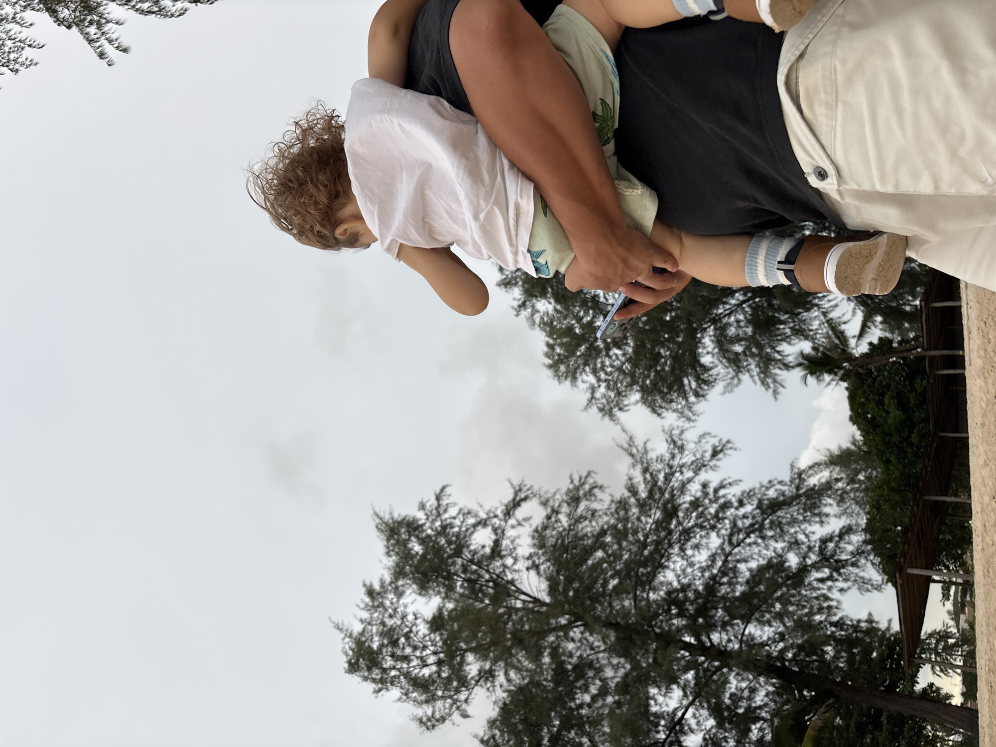 A person holds a small child in their arms. The child, with curly brown hair, is wearing a white t-shirt and patterned shorts. The person is wearing a dark t-shirt and light-colored pants. The child's feet are visible, wearing striped socks and brown shoes. The person is holding a smartphone in their hand.
The scene is outdoors, with trees and a cloudy sky visible. The overall impression is casual and relaxed. The time of day appears to be daytime, given the light, and the weather seems overcast. The location context provided is Choeng Thale, Thailand, which suggests a tropical or subtropical environment. There is no visible text in the image.