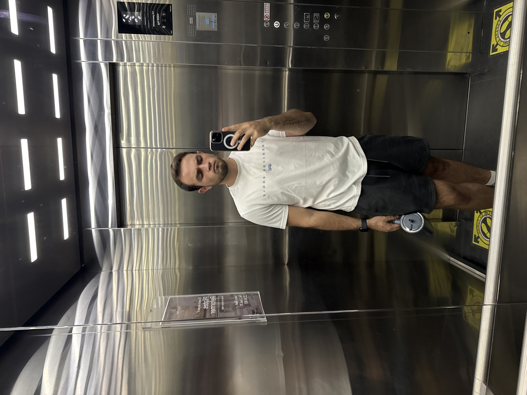 The image captures a man taking a selfie inside a modern elevator with highly reflective metallic walls. The man, with short dark hair and a beard, wears a white t-shirt with the mirrored text "WE MEDICIN EUROPE" and dark shorts. He holds a black smartphone, featuring a visible magnetic ring, in his left hand, angling it to capture his reflection. In his right hand, he holds a silver water bottle or thermos. His expression is neutral.
The elevator's interior is dominated by polished stainless steel, creating numerous reflections of the man, the phone, and the horizontal strip lights visible on the opposite wall. The elevator control panel, located on the right wall from the man's perspective, is clearly visible. Its digital display shows "L" (likely for lobby) and the capacity: "1000 kg" and "13 personas" (13 people). Mirrored text on the panel includes "OTIS" and "PELIGRO: NO USAR EL ASCENSOR EN CASO DE INCENDIO" (Danger: Do not use the elevator in case of fire).
Near the bottom of the right wall, a yellow warning sign, also seen in reflection, features a circular symbol with a diagonal line through it and mirrored text that reads "NO TOQUE LA PUERTA" (Do not touch the door). An information panel, suspended lower down on the left wall, shows mirrored text, partially readable as "Pilates Reformer". The visible Spanish text aligns with the provided location context of San Bernardino Tlaxcalancingo, Mexico.