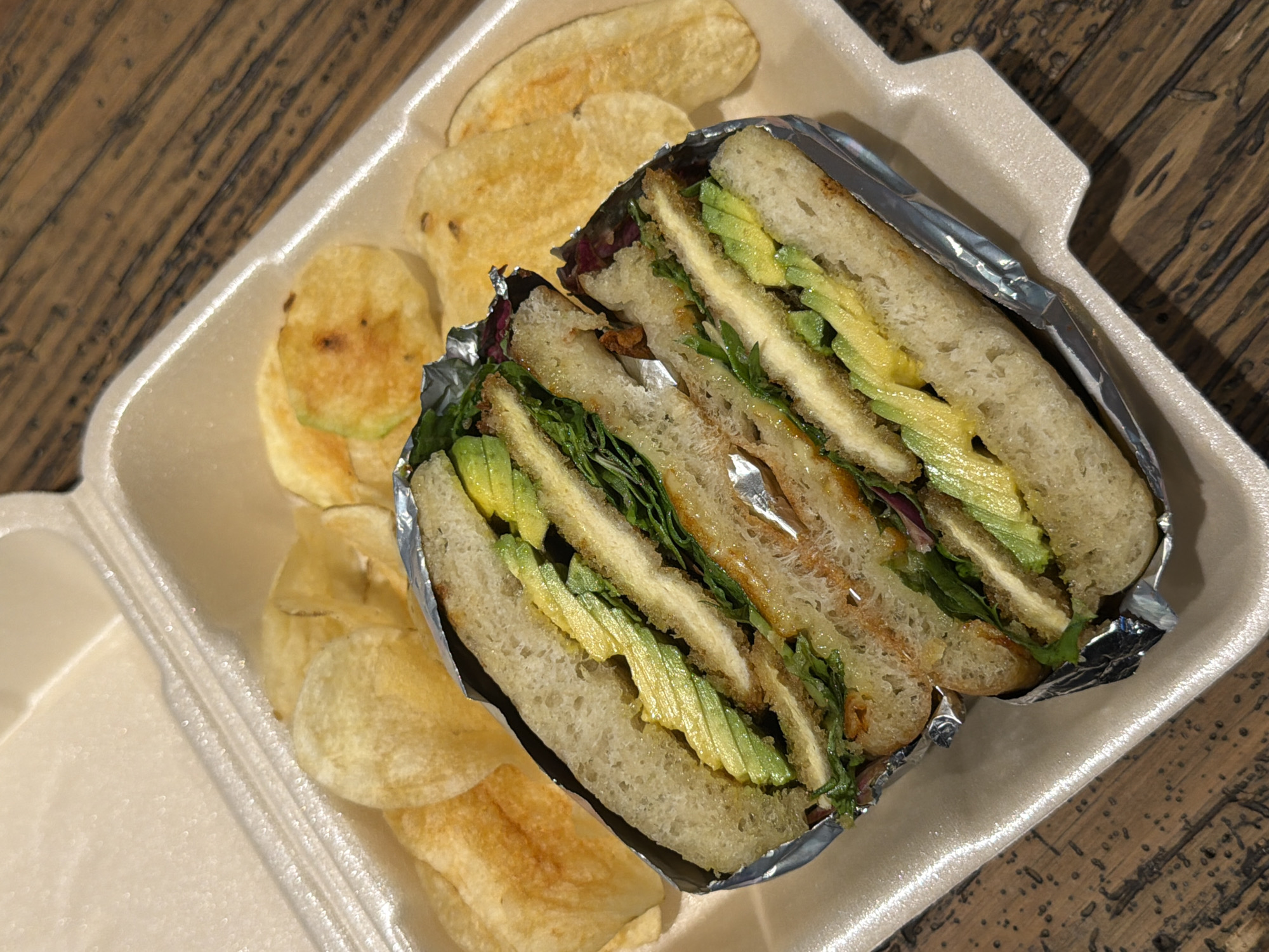 A sandwich cut in half and wrapped in aluminum foil rests in a white styrofoam container alongside several potato chips. The container sits on a wooden table. There is no information available to determine if the photograph was taken in Puebla, Mexico or any other location. No people, actions, or interactions are visible in the image.