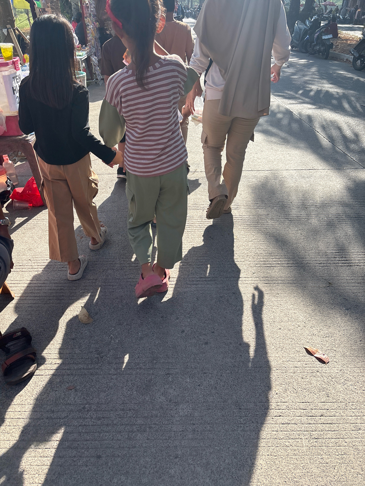 In Telagamurni, Indonesia, a street scene unfolds with three individuals walking on a concrete path. Two young girls, one in a black long-sleeved shirt and tan cargo pants, the other in a horizontally striped long-sleeved shirt and green pants, hold hands as they walk away from the viewer. Their footwear includes white sandals for the girl in black and pink slides for the girl in stripes. To their right, an adult, seen from the back, wears a light-colored top, tan cargo pants, and beige sandals. The adult is partially obscured by a draped beige garment. The environment features a sidewalk lined with trees casting dappled shadows on the ground. In the background, stalls with various goods, including a drink dispenser with "TEH HI 2" visible, are set up. Motorcycles are parked along the side of the road. The ground is marked with horizontal lines, indicating a paved surface. A single brown leaf lies on the concrete in the foreground. The lighting suggests it is daytime, with strong shadows indicating sunlight.