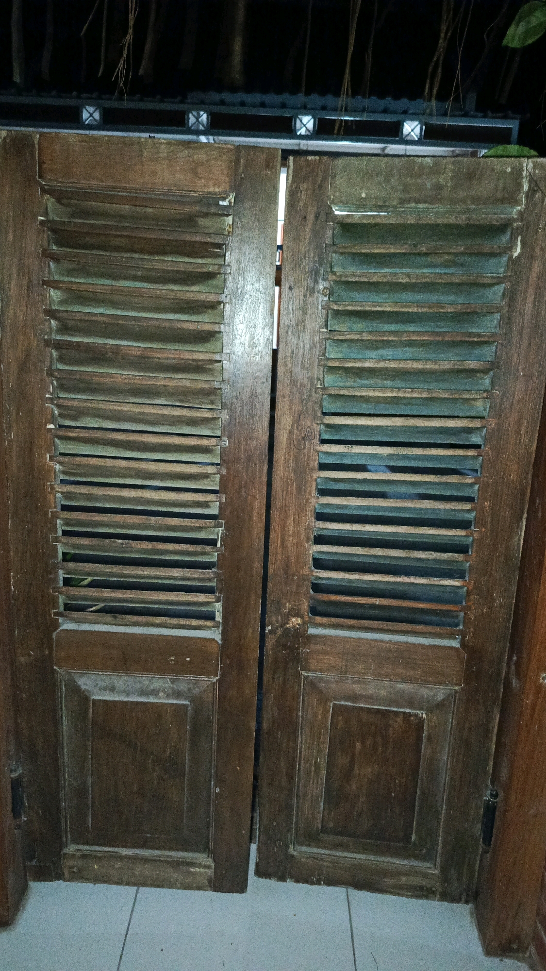 The image displays a close-up, head-on view of two weathered, louvered wooden doors. The doors are made of dark wood, with horizontal slats creating an open, ventilated design. The bottom portion of each door features a raised, rectangular panel with beveled edges. The wood shows signs of age and wear, with a dusty, aged patina. In the background, obscured by the doors, a dark, possibly covered area with hanging vines is visible. The doors are standing upright, leaning slightly, and appear to be at ground level, with a light-colored tiled floor visible at the bottom. There are no people or animals present. The scene appears to be outdoors, possibly in a backyard or patio area. The overall impression is one of aged, functional, outdoor furniture. The location in Babadan, Indonesia, suggests a tropical or subtropical climate, which aligns with the presence of vines.