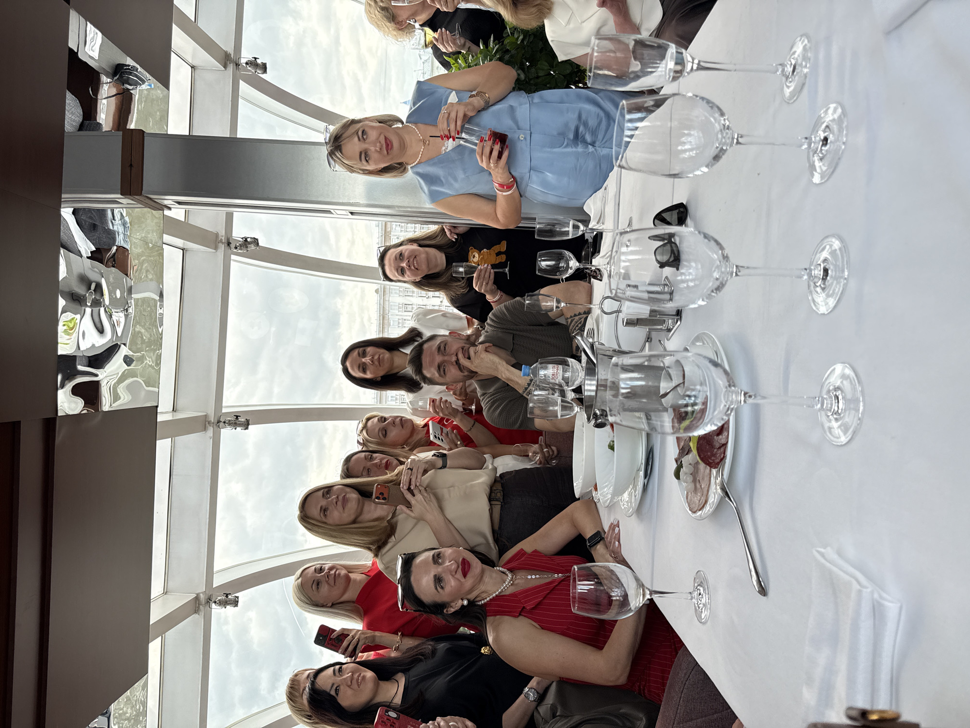 A group of eleven people, predominantly women, are seated at a long table covered with a white tablecloth, inside what appears to be a restaurant or event space with large windows. Several of the women are holding smartphones, possibly taking photos. The table is set with wine glasses, plates, and cutlery. One plate has a selection of cured meats on it. A bottle of water is also visible. The background shows a cityscape through the windows, with a cloudy sky. The location is Moscow, Russia.