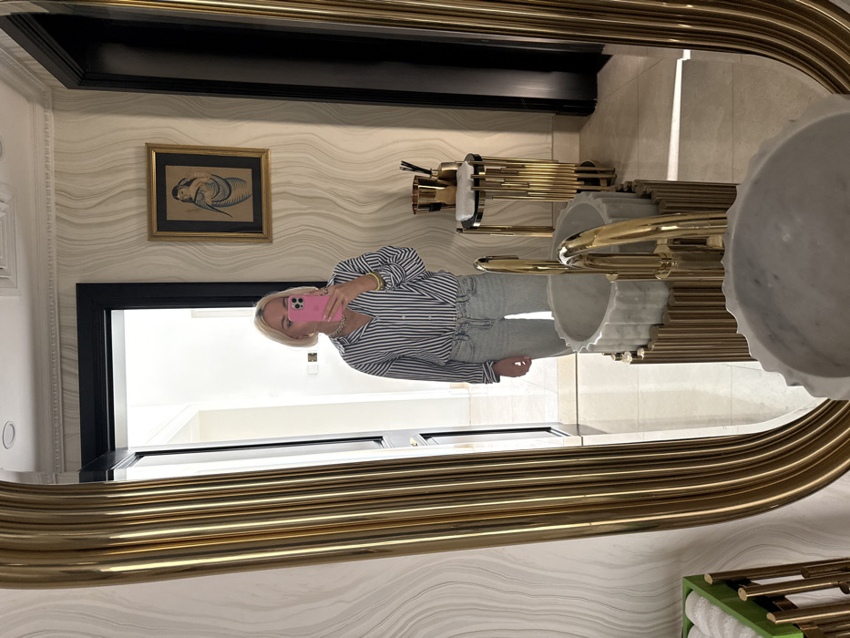 The image depicts a mirror selfie taken by a blonde individual, identified as female, within a luxurious interior space. The subject wears a blue and white striped long-sleeve shirt, light-wash denim jeans, a gold chain necklace, gold hoop earrings, a bracelet, and rings, holding a pink smartphone to capture the reflection.
The mirror itself is large and oval-shaped, featuring a thick, fluted gold-colored frame. Reflected prominently are two round, fluted white marble sinks with curved gold faucets, each resting on a cylindrical base composed of vertical gold rods. The background wall is covered with light-colored wallpaper displaying a subtle wavy pattern. A framed artwork, depicting a stylized figure in monochromatic tones within a gold frame, hangs on this wall. To the right of the sinks, a slender stand with a gold-rod base and circular top holds a folded white towel and multiple gold-colored containers. A dark-framed doorway is visible, leading to a brightly lit interior space with a light-colored tiled floor. A green and gold stand holding folded white towels is partially visible on the far left. The overall environment, characterized by opulent fixtures, marble, and gold accents, is consistent with high-end luxury interiors found in the city of Dubai, United Arab Emirates, likely within a private residence or a luxury hotel bathroom/powder room.