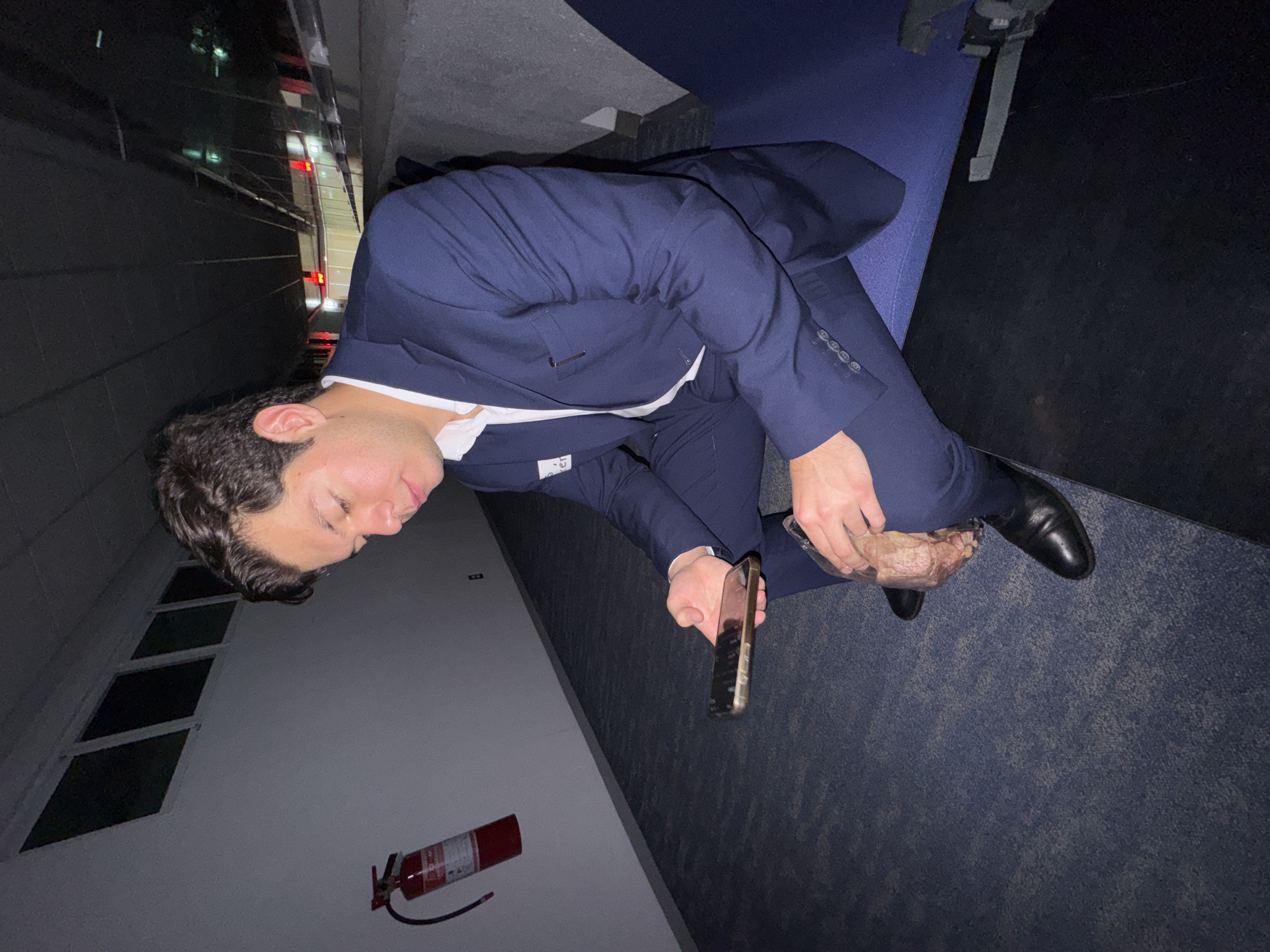 A man wearing a navy blue suit with a white shirt is sitting on a blue surface. He is holding a phone in his left hand and a clear plastic container of meat in his right hand. The background shows an exterior wall with a fire extinguisher mounted on it and what appears to be the skyline of Puebla, Mexico.