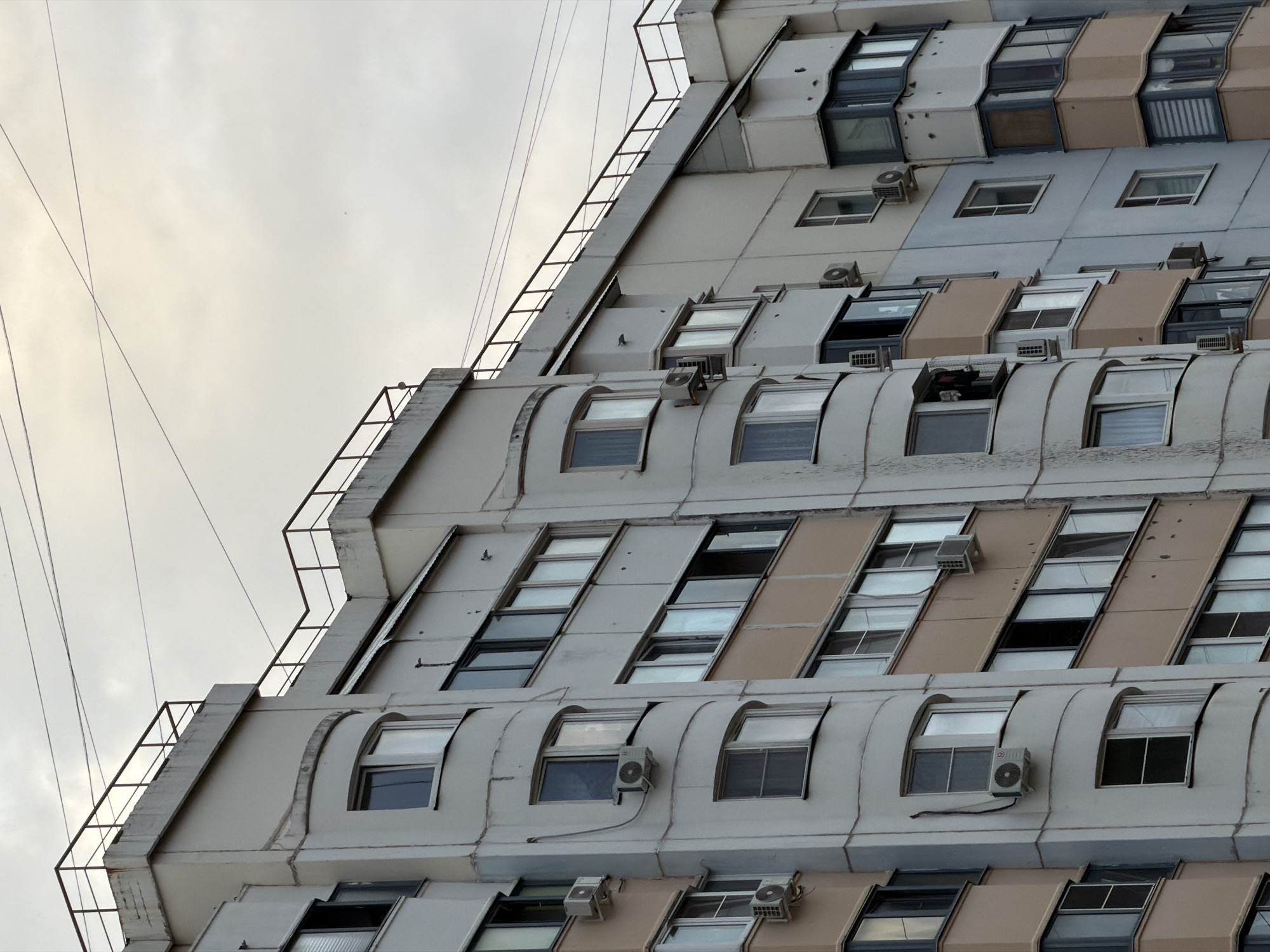 The image shows the exterior of a multi-story apartment building in the city of Khimki, Russia. The building is constructed with light-colored concrete and has a series of balconies and windows. Several air conditioning units are visible mounted on the exterior walls. The sky is overcast, and power lines are strung across the top of the frame. The architectural style appears to be typical of Soviet-era mass housing. No people are visible in the image.