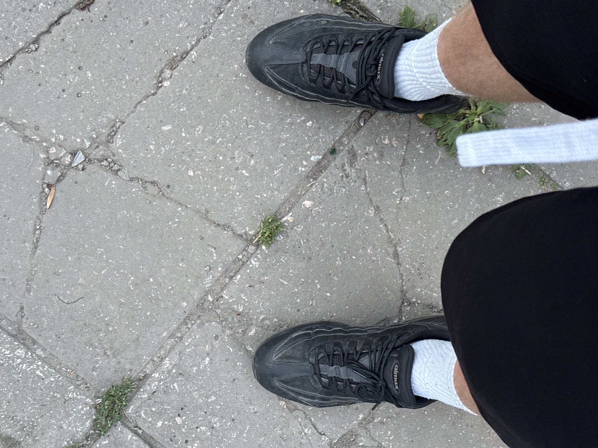 A first-person perspective image shows a person's feet wearing black athletic shoes and light grey socks, standing on a cracked concrete sidewalk. Patches of grass and small debris are visible between the concrete slabs. The scene is likely outdoors, with natural lighting. The location is identified as Zaporizhzhia, Ukraine. A white object, possibly a piece of paper or fabric, is visible near the right foot.