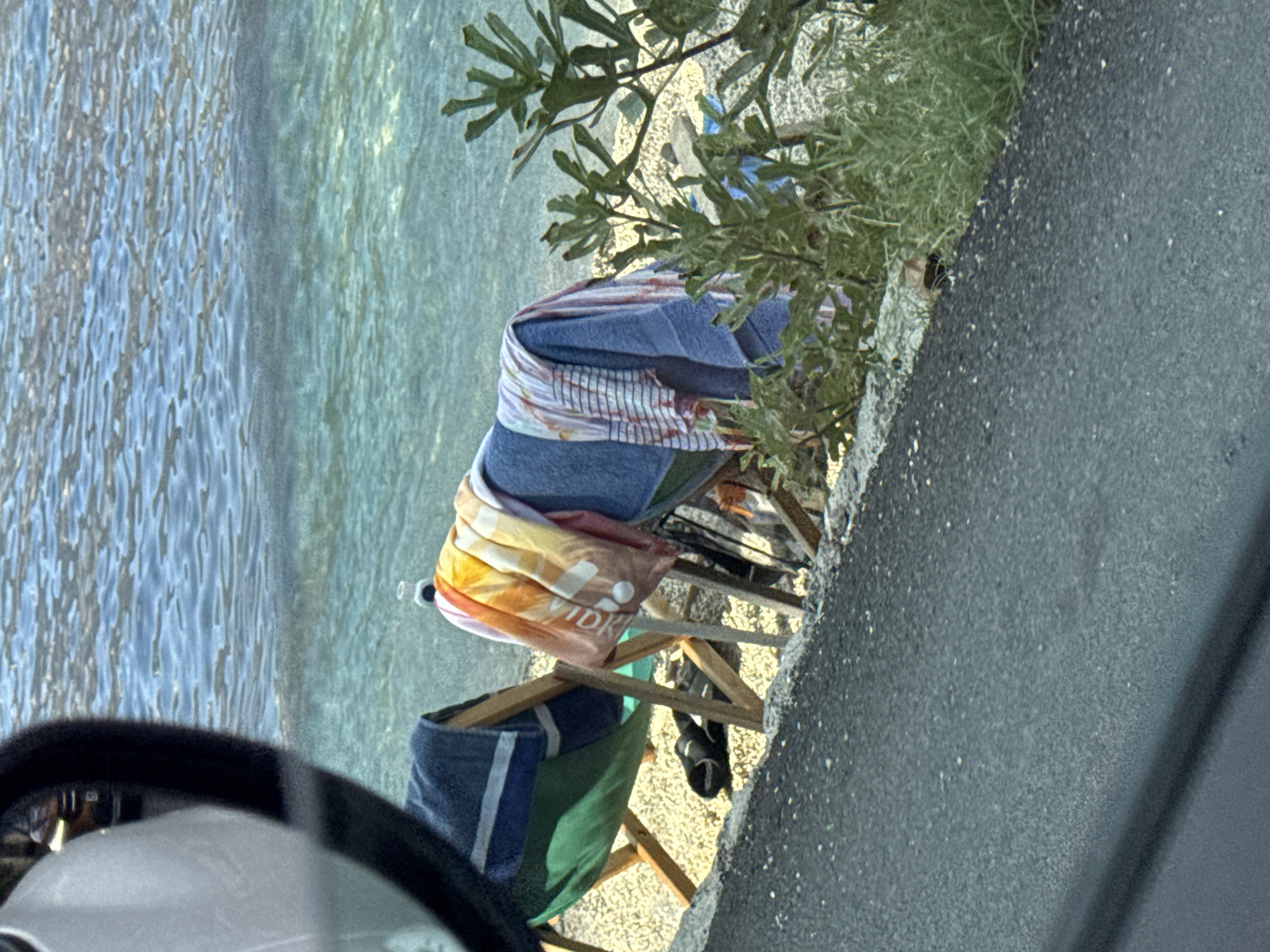 The image captures a sunlit scene near the water, likely a beach or waterfront area in Prčanj, Montenegro. The foreground features a portion of a vehicle's interior, specifically a side-view mirror, reflecting a bright, outdoor environment. This suggests the photo was taken from inside a car parked near the water.

The main subjects are several beach towels, casually folded or draped over a wooden beach chair. One towel displays a vibrant pattern of orange, yellow, and white stripes, with visible text that appears to read "VIDKI". Another towel is a solid blue with white stripes, and a green towel is also present. The beach chair is made of light-colored wood.

Behind the chair, lush green foliage is visible, hinting at a vegetated area adjacent to the beach. The water is clear and blue, with visible ripples indicating a gentle movement. The ground next to the vegetation is a rough, grey asphalt surface. The overall impression is one of a relaxed, outdoor setting, possibly a day at the beach or a stop by the coast. The lighting suggests it is daytime, with bright sunlight casting clear shadows and illuminating the scene. The presence of beach towels and the proximity to the water strongly indicate a recreational context, possibly a break from driving or a moment of leisure.