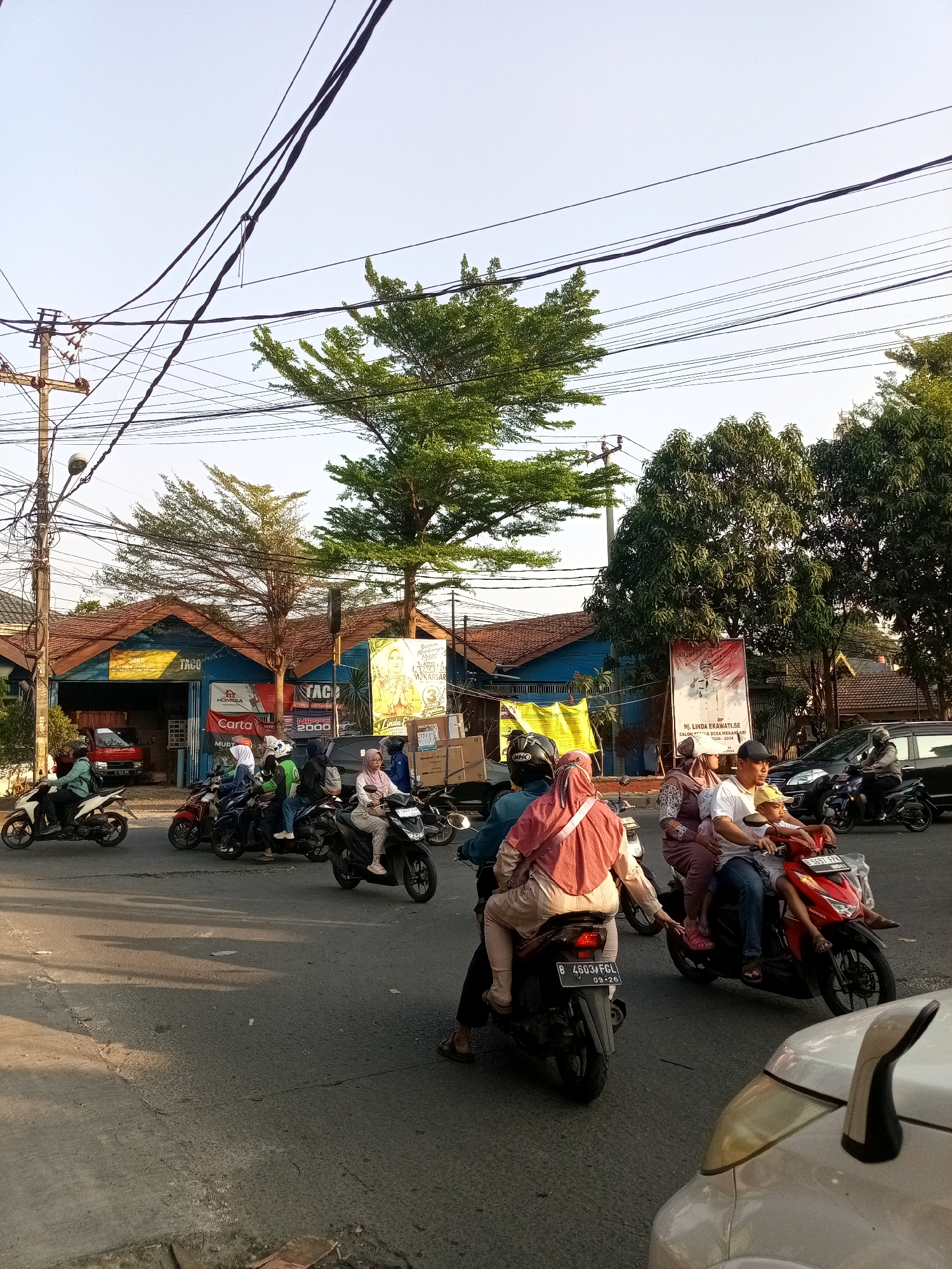 In Tambun, Indonesia, a street scene unfolds with several motorcycles carrying passengers navigating a road. A red Honda motorcycle, with license plate B 5657 FPA, carries a driver and two passengers, one a child wearing a yellow cap and the other an adult in a white shirt and blue jeans. To their left, a black motorcycle, license plate B 4603 FGL, carries two individuals, one wearing a pink hijab and the other a tan-colored outfit. Another motorcycle, this one white and driven by a person in a light blue shirt, is positioned further left. In the background, several buildings with brown tiled roofs are visible, some displaying colorful banners. One banner features an image of a woman and text, likely promotional. Trees with vibrant green foliage are interspersed with the buildings and line the street. Overhead, a complex network of power lines stretches across the sky, supported by utility poles. The road surface appears to be asphalt, with visible cracks and debris. The overall atmosphere suggests a typical day with local transportation and commercial activity.