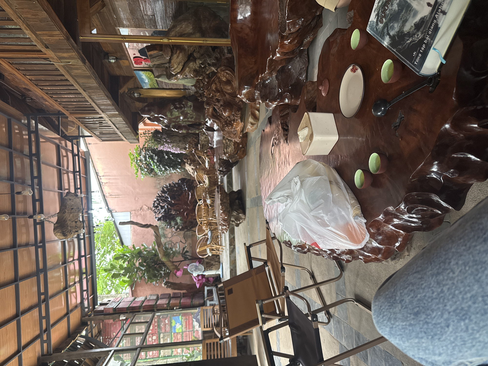 The image depicts a scene in Bản Đêu, Vietnam, likely within an outdoor or partially covered dining area. A large, intricately carved wooden table dominates the foreground, featuring items such as teacups, a spoon, a book, a plate, a tissue box, and a plastic bag. A person wearing blue jeans sits beside the table. Several chairs are arranged around the table. Beyond the table, the area includes a garden with various plants and flowers. A building or structure with glass windows and a metal frame is visible.