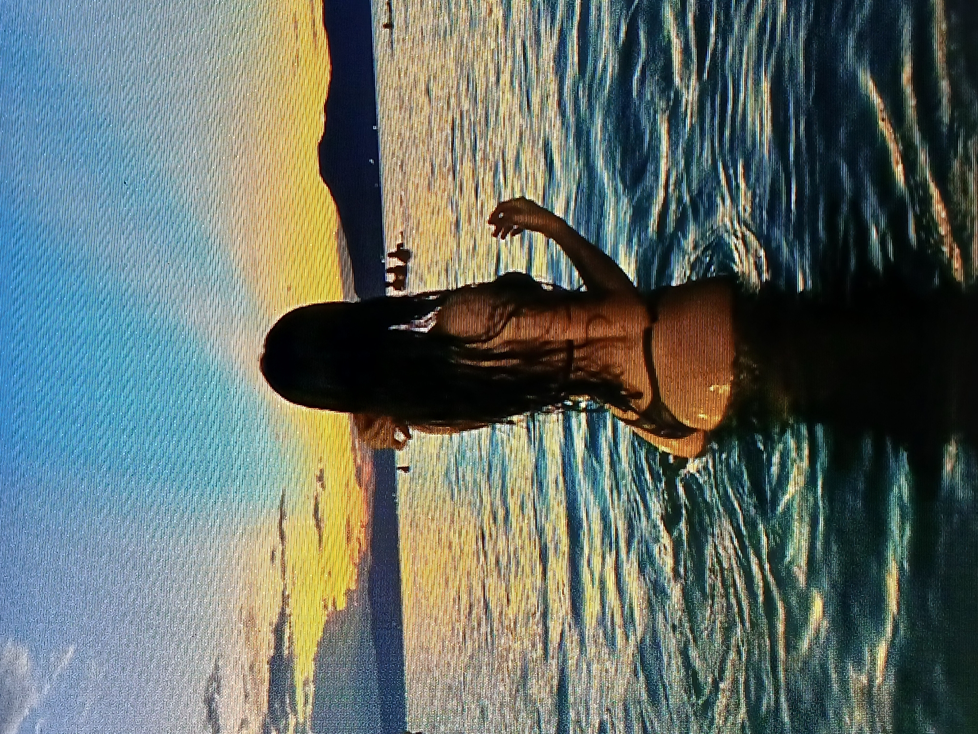 A woman with long dark hair is wading in the ocean at sunset, likely near Juan Griego, Venezuela. The water appears to be relatively calm with subtle waves reflecting the golden light of the setting sun. The sky transitions from blue to orange as the sun dips towards the horizon. The land is visible in the distance.