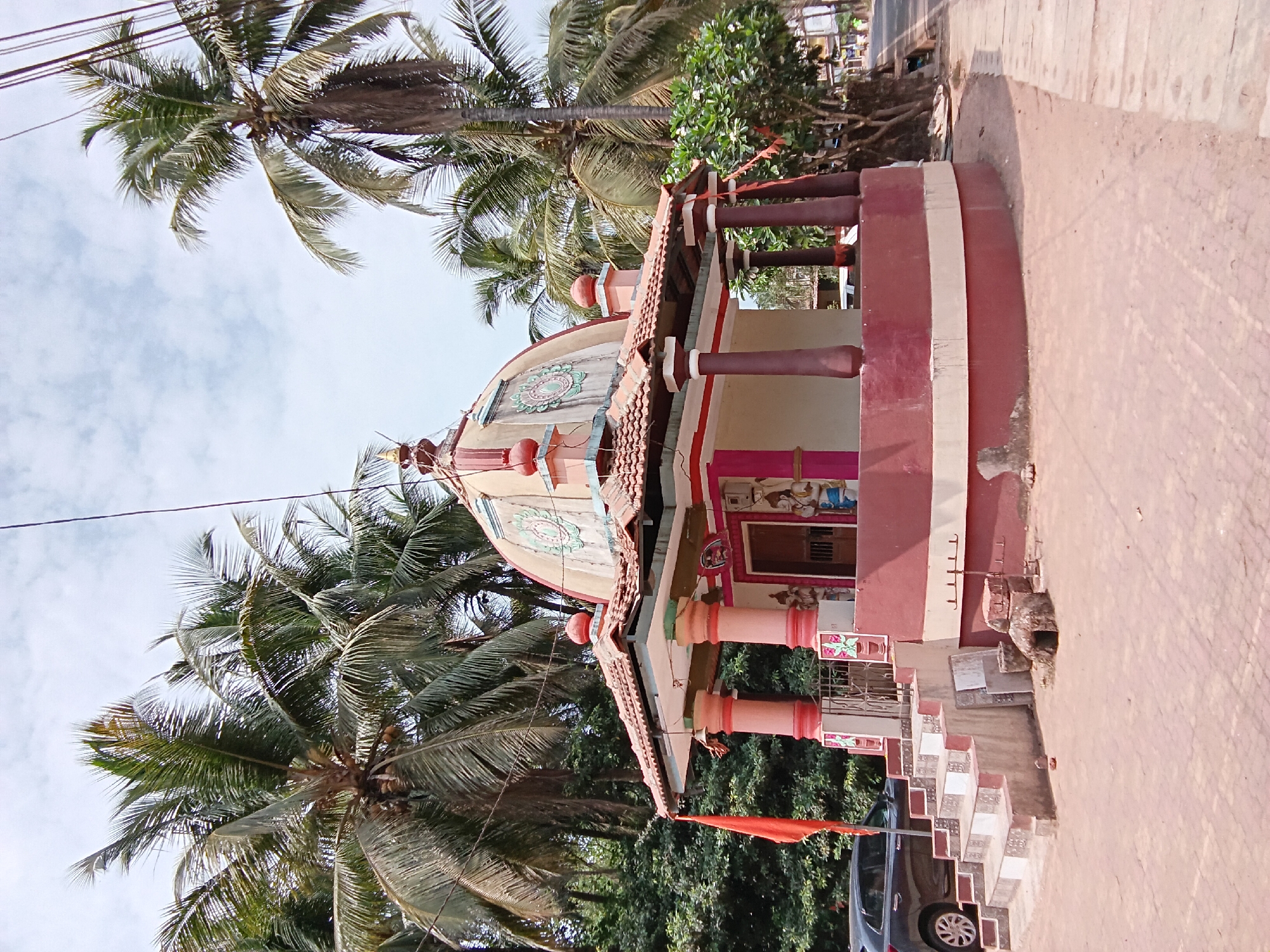 The image depicts a small, colorful Hindu temple in Chopdem, India, situated amidst palm trees. The temple has a dome-shaped roof adorned with decorative patterns and a red flag flying nearby. The structure is painted in shades of red, white, and pink, with pillars and a railing surrounding the main shrine. A paved pathway leads to the temple entrance. A car is visible parked on the side of the road adjacent to the temple steps. The sky is overcast, suggesting a cloudy day.